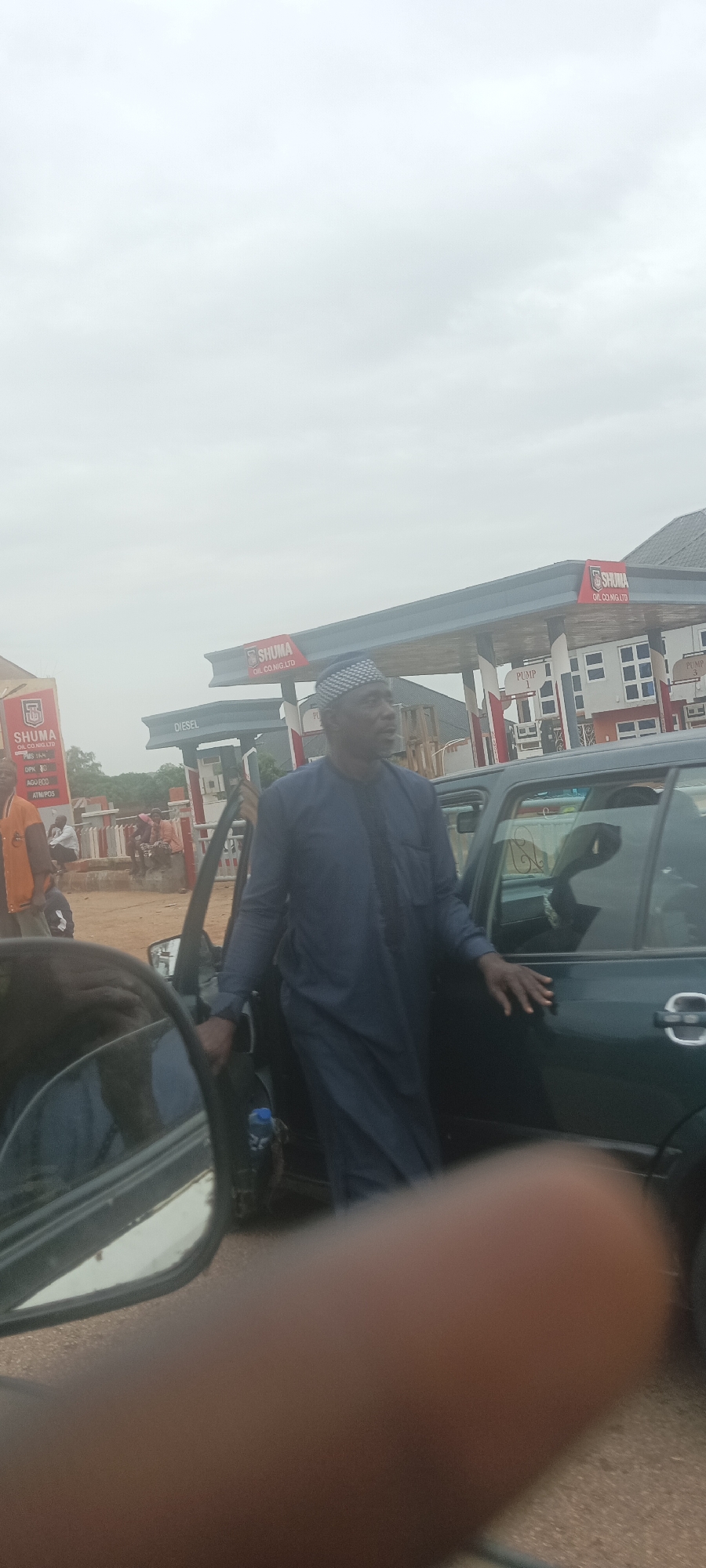 The image captures a scene at a Shuma Oil Co. Nig. Ltd gas station. A man wearing a dark blue traditional outfit and a patterned cap is standing next to a dark green car, with the car door open. A finger is visible in the foreground, obscuring some of the view. The gas station has pumps labeled "DIESEL" and "PUMP 1," and a sign indicating ATM/POS availability. People are visible in the background, some sitting and others on a motorcycle. The sky is overcast. The location appears to be in Durimi, Nigeria, based on the gas station signage.