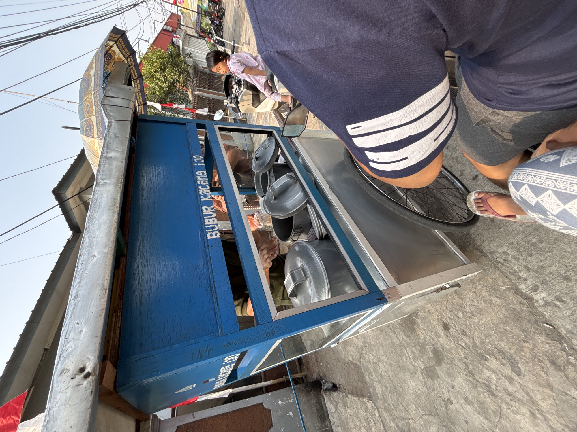 This is a medium-shot, eye-level, outdoor photograph taken in Bekasi, Indonesia, showing a street food cart on a concrete sidewalk. The cart is primarily blue and silver, with a large blue wooden front panel displaying white text that reads "BUBUR KACANG IJO". A large, patterned umbrella is open and propped on top of the cart. Several metal pots and lids are visible inside the cart, with a person's hands in the background, appearing to be preparing food. A young girl in a pink outfit is sitting on a motorcycle parked next to the cart, looking to the side. In the foreground, the backs of two people are visible, one wearing a dark blue shirt with white stripes and the other wearing patterned shorts and flip-flops. The environment includes buildings and utility wires in the background, suggesting an urban setting. The lighting indicates it is daytime.