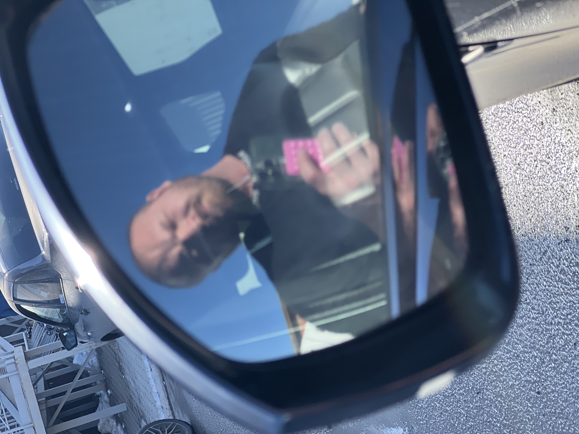 The image depicts an outdoor scene in Chaikovsky, Russia, captured as a reflection in a vehicle's exterior side mirror. The mirror's dark frame partially borders the reflection. Within the reflection, an adult male with a bald head and a beard is visible, wearing a dark-colored shirt and a silver-colored chain around his neck. He is holding a pink smartphone in his right hand, directed towards the mirror, suggesting a self-photograph or video recording. The background visible outside the reflection includes a dark-colored sedan, identified by a "GRANTA CLUB" sticker on its rear window, parked on a paved surface. Behind the sedan, a white, multi-level structure, potentially scaffolding or a small building, stands over an area with accumulated snow or debris. The immediate foreground and areas beneath the vehicles consist of wet, granular asphalt. A partial view of a car wheel is present on the far left. The scene is illuminated by daylight under clear sky conditions, reflected in the mirror's blue hue.
