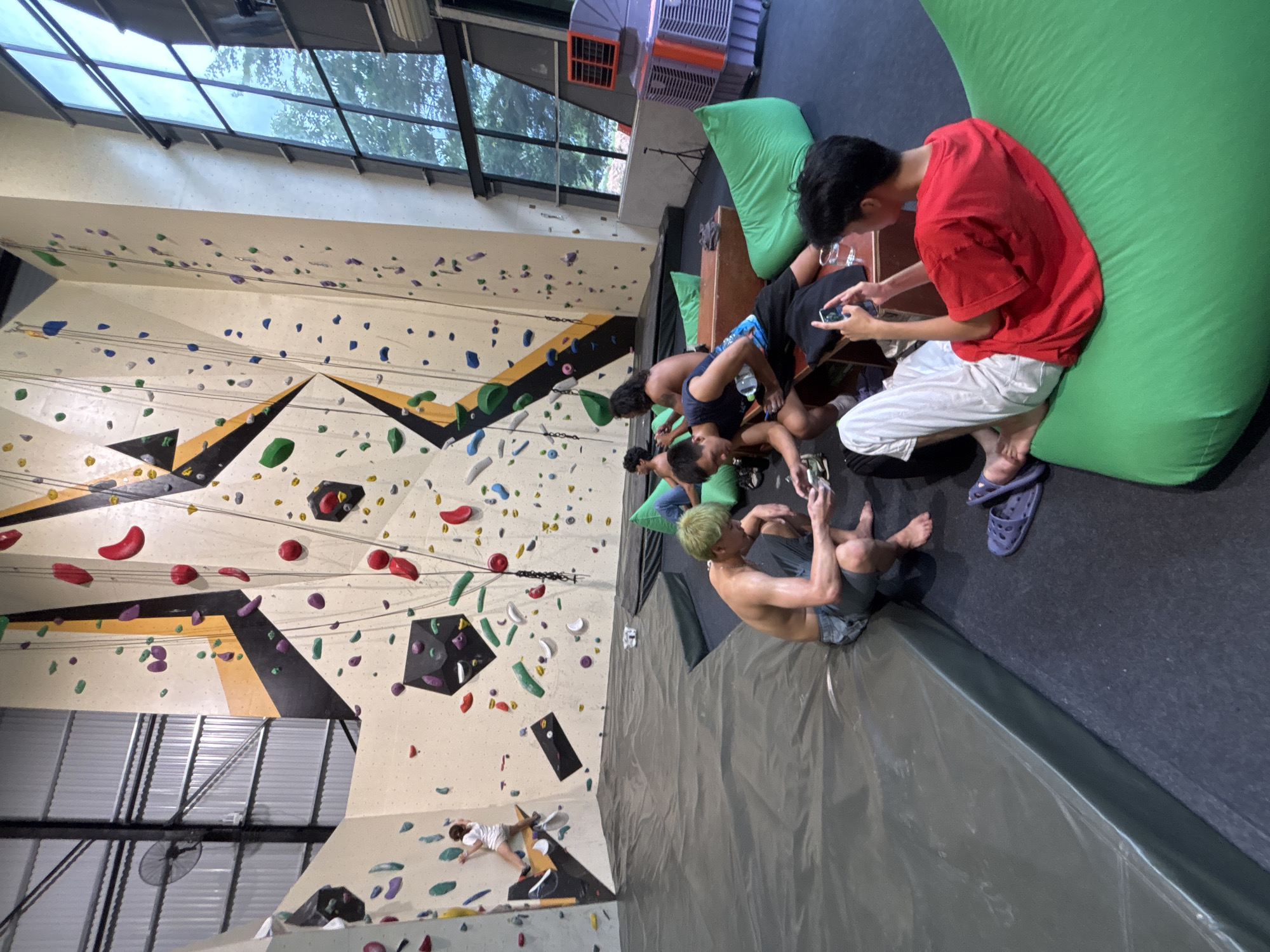 The image depicts the interior of a climbing gym, likely located in Đà Nẵng, Vietnam, characterized by its high walls adorned with colorful climbing holds and ropes. Several individuals are present in the gym, with a focus on a group resting on the floor amongst large, green beanbag chairs and wooden benches.
In the foreground, a young man with green-dyed hair is seated on the mat, wearing grey shorts and shirtless. He is looking down at his hands, which are holding a mobile phone. To his right, another young man, shirtless and wearing grey shorts, is also seated and appears to be engaged with a phone. Further to the right, a young man in a red t-shirt and white pants is seated on a large green beanbag chair, holding a phone. Another person, partially visible, is seated on a bench next to him. In the background, on the climbing wall, a lone individual is climbing. The gym environment includes a large fan mounted on the corrugated metal ceiling and extensive windows revealing trees and the sky outside. The overall atmosphere suggests a relaxed break or post-climbing session.