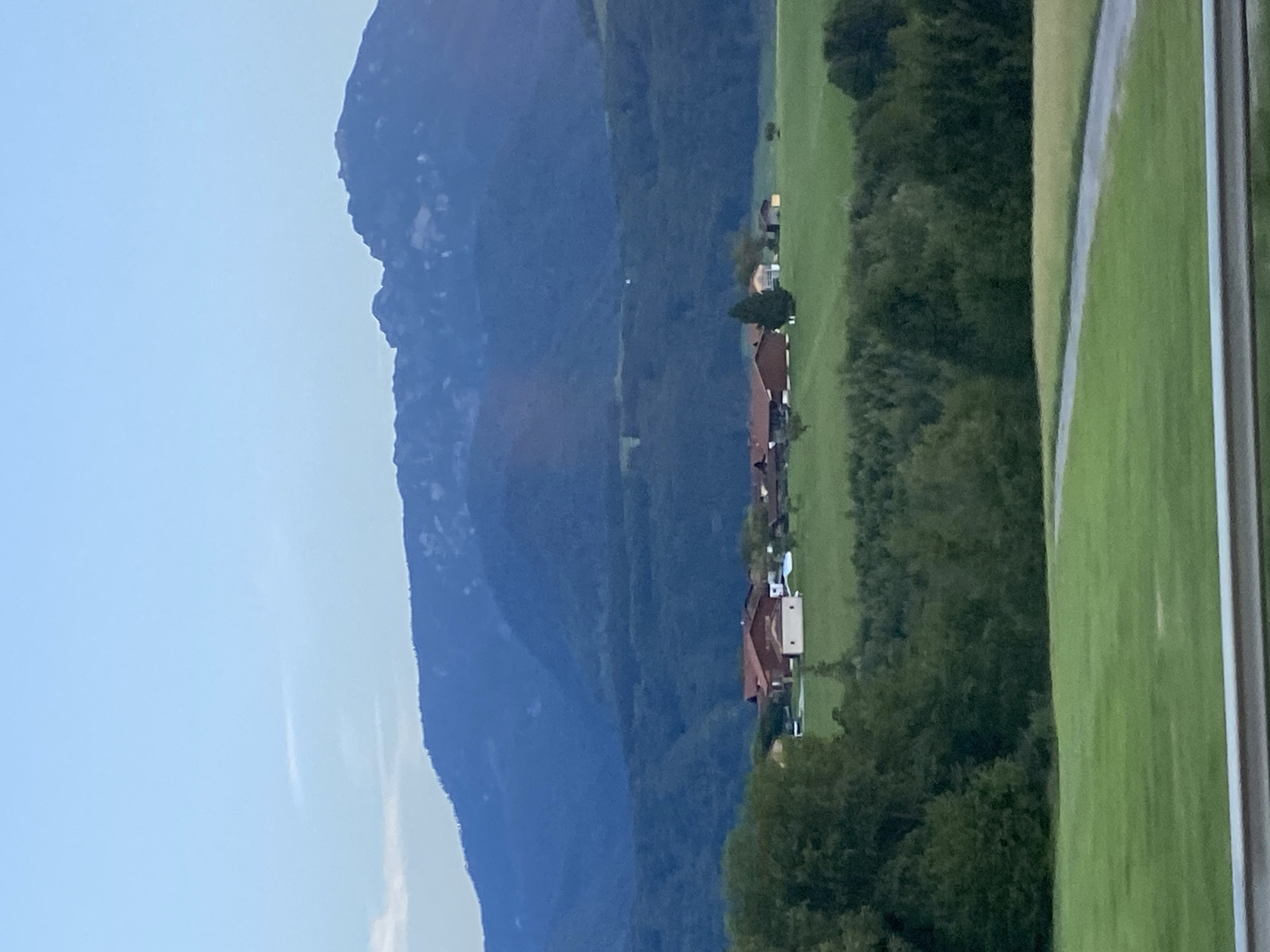 This is a vertical, eye-level, medium shot photograph taken from a distance of a rural landscape in the city of Bergen, Germany. In the foreground, a portion of a metallic guardrail is visible. Beyond the guardrail is a sloped green field. A grey paved path winds its way through the field. In the midground, a collection of buildings with red roofs, characteristic of a farm or village, are situated on a green hillside. A few trees are interspersed among the buildings. Further back, a dense forest blankets the rolling hills. In the background, a large, imposing mountain range with a craggy peak dominates the horizon, its slopes shrouded in deep blue hues. The sky above is a pale, clear blue with a hint of a thin, wispy cloud. The overall scene depicts a serene, natural landscape with human settlement nestled within.