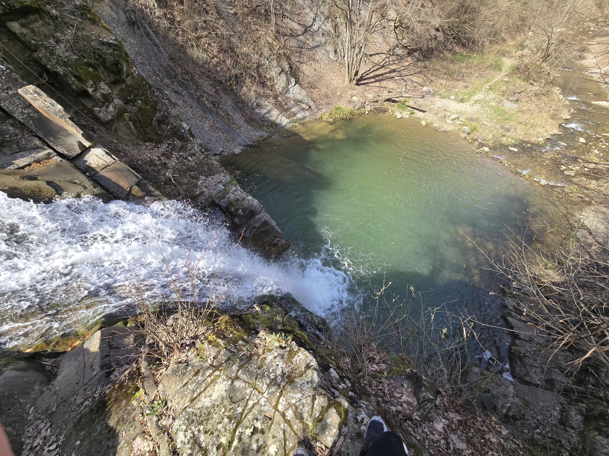 A high-angle shot depicts a waterfall cascading into a small, clear lake. The falls are framed by rock formations on the left, while the lake is bordered by trees and a stream on the right. The presence of water and vegetation suggests a natural environment. Part of a person's legs and feet are visible in the foreground.