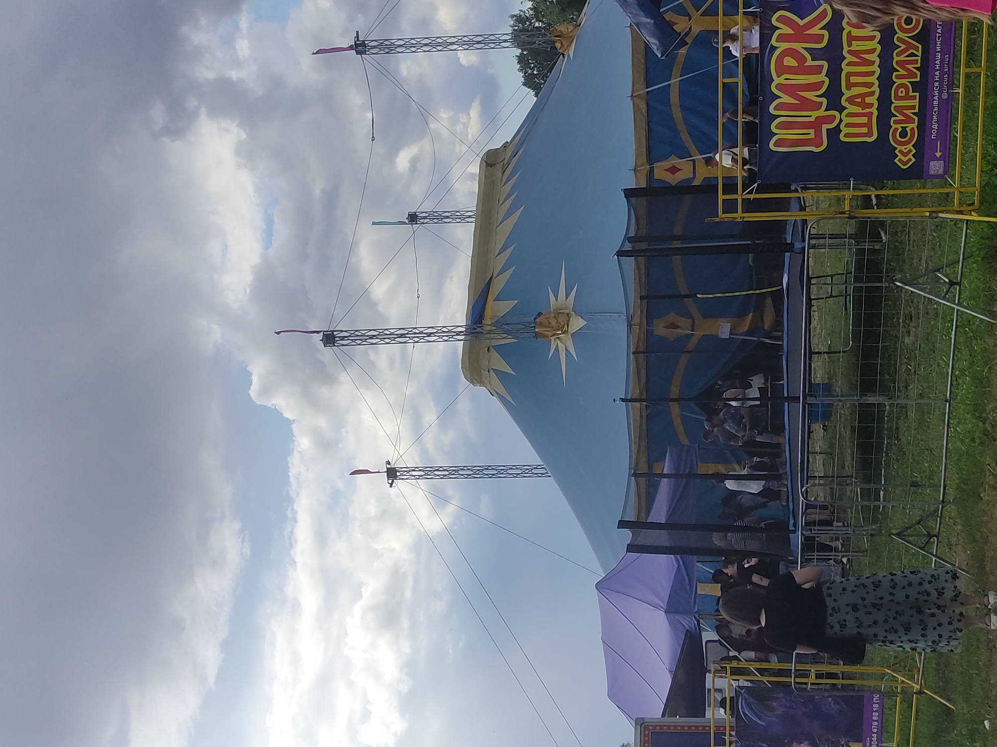 The image depicts a circus tent and surrounding area, likely in Mahilyow, Belarus, under a cloudy sky. A large blue and yellow striped circus tent with a pointed roof dominates the center of the frame. Several tall, metal poles support the tent with wires extending between them. In the foreground, a trampoline enclosed by a black net is visible, with several people standing or walking around it. To the left of the trampoline, a smaller purple tent is set up. A woman in a black top and a patterned skirt stands facing away from the camera, looking towards the trampoline area. A yellow barrier with a blue and yellow sign advertising "ЦИРК ШАПИТО <<СИРИУС>>" (Circus of tents "Sirius") is positioned to the right of the trampoline. The ground is covered in grass, and trees are visible in the background.