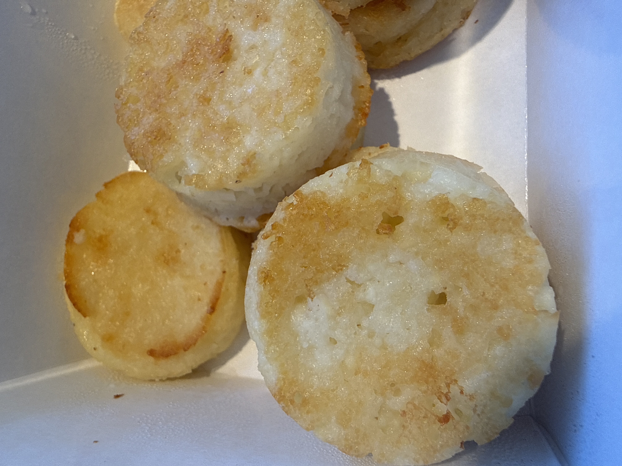 The image shows a close-up of several small, round, golden-brown fried potato pancakes stacked and layered in a white cardboard container. The pancakes have a slightly irregular, textured surface with some crispy edges. The lighting suggests the food is indoors, possibly in a restaurant or take-out setting. There is no visible indication of the specific location within Orenburg, Russia.