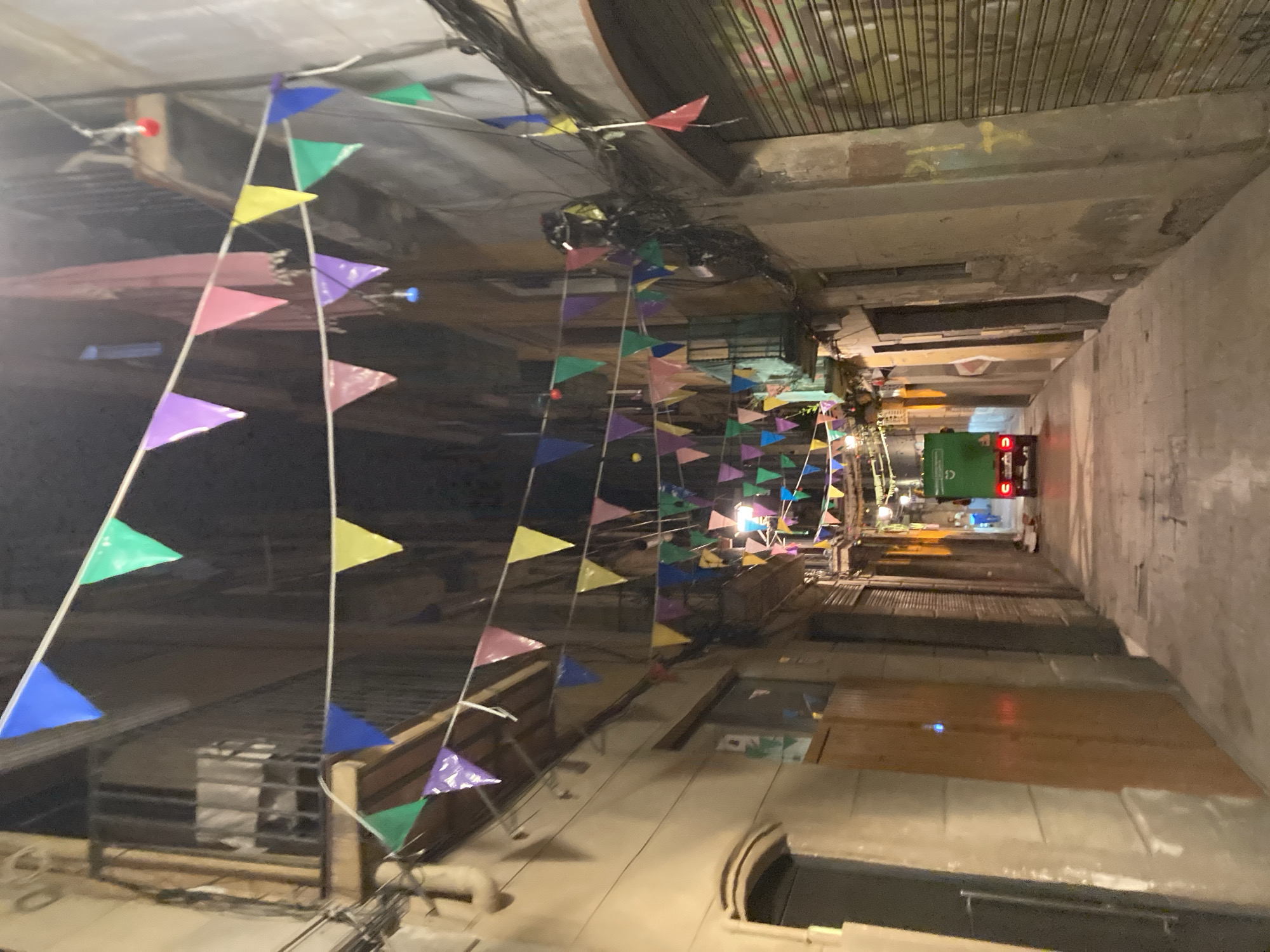 The image depicts a narrow, illuminated alleyway in Barcelona, Spain, adorned with strings of colorful triangular flags. The flags, in hues of blue, green, yellow, pink, and purple, stretch across the alley, suspended between the aged buildings that flank it. These structures, with their balconies and window frames, suggest a historic urban setting.
At the far end of the alley, a bright green motorized vehicle, possibly a small truck or cart, is visible, with its taillights illuminated. The vehicle is positioned on the paved pathway of the alley. The ground is a concrete surface, showing signs of wear. Overhead, a dense network of electrical wires crisscrosses the space, adding to the urban feel. The lighting in the alley appears to be a combination of ambient light from the buildings and the vehicle's headlights, along with some overhead string lights or festive illuminations. The overall atmosphere is that of a vibrant, possibly festive, street at night.