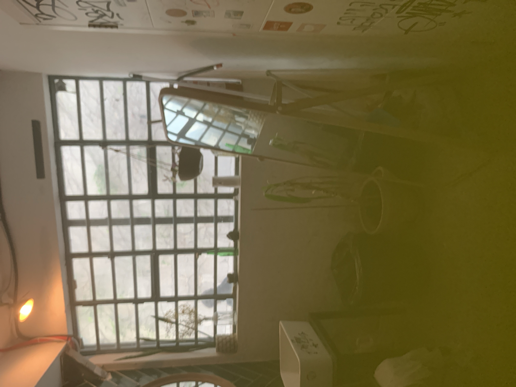 A room in Tel-Aviv, Occupied Palestine, features a multi-pane window with a view of the outdoors. A mirror is propped against the window frame. A sink, various containers, and objects are arranged near the window. The walls are decorated with writing. A light fixture is mounted on the wall.