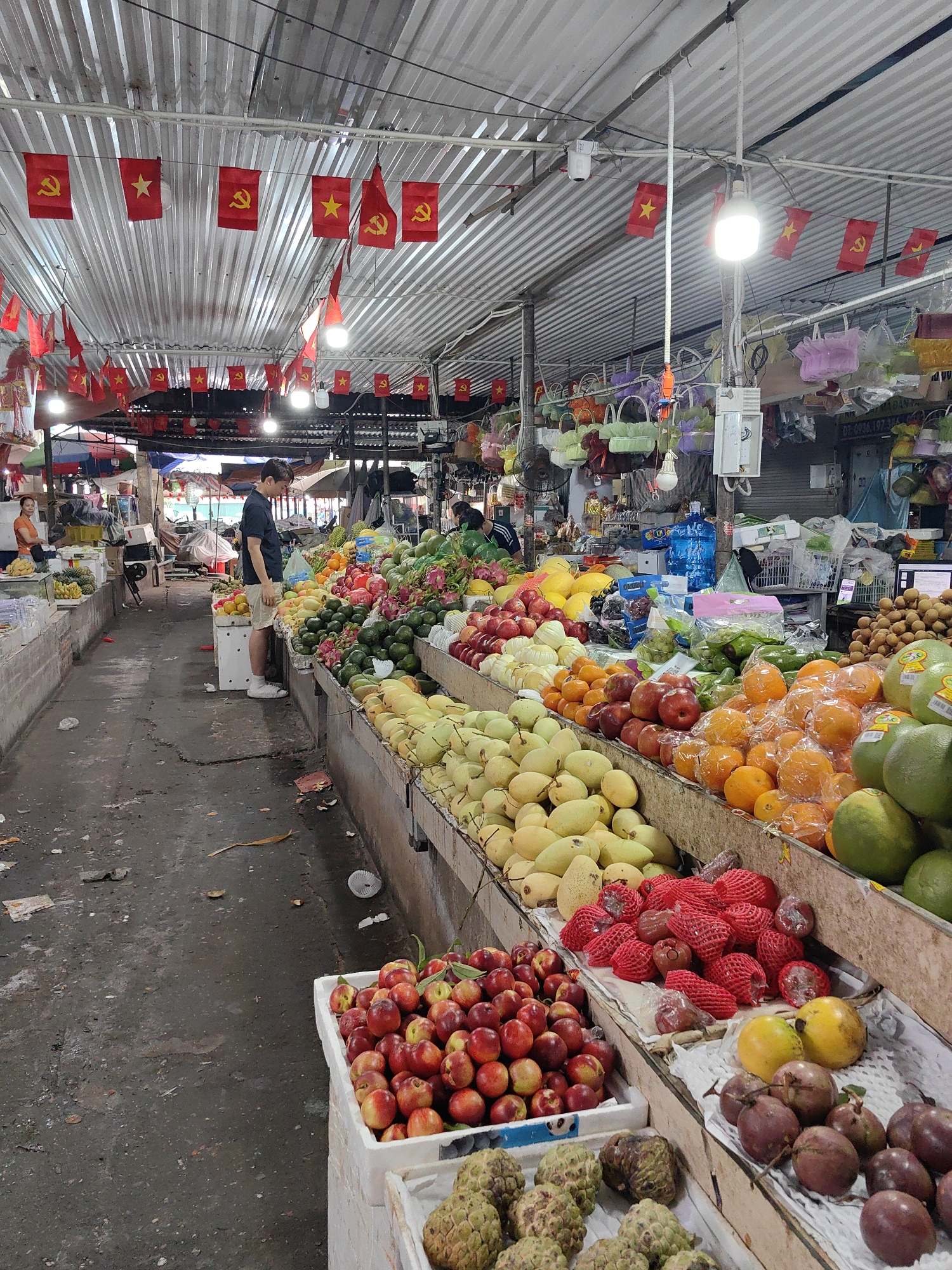 The scene depicts a bustling indoor market in Bai Chay Ward, Vietnam, filled with a variety of fruits displayed along concrete stalls. Overhead, numerous red flags with a yellow star and sickle hang from the corrugated metal roof, typical symbols associated with the Vietnamese Communist Party.
The market's concrete floor is stained and littered with debris. A man stands in the aisle, appearing to examine the fruit on display. The stalls overflow with colorful produce, including mangoes, oranges, dragon fruit, and other tropical fruits. Some of the fruits are packaged in red netting.
Fluorescent lights illuminate the scene, casting harsh shadows throughout the market. In the background, hints of other stalls and vendors can be seen.