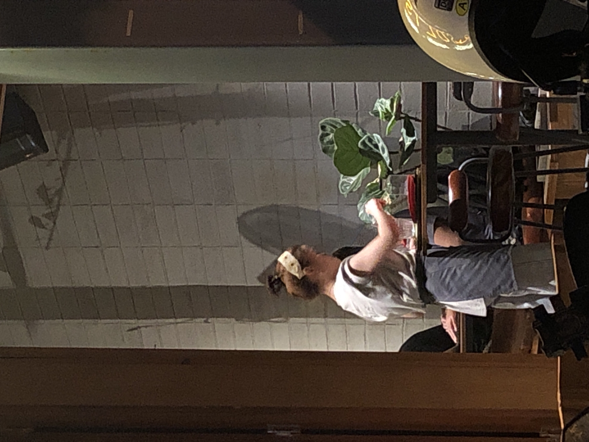 The image depicts a low-angle shot of a person sitting at a table. The person is wearing a white t-shirt and a light-colored headband. They are holding a clear glass with a drink in their right hand. Their left arm is bent and they are reaching out to a plant with large, green leaves to their left. The setting appears to be indoors, with a tiled wall in the background. Part of a light fixture with visible text is visible in the upper right corner. The overall lighting suggests it could be evening or indoors with artificial lighting. The scene is set in Da Nang, Vietnam.