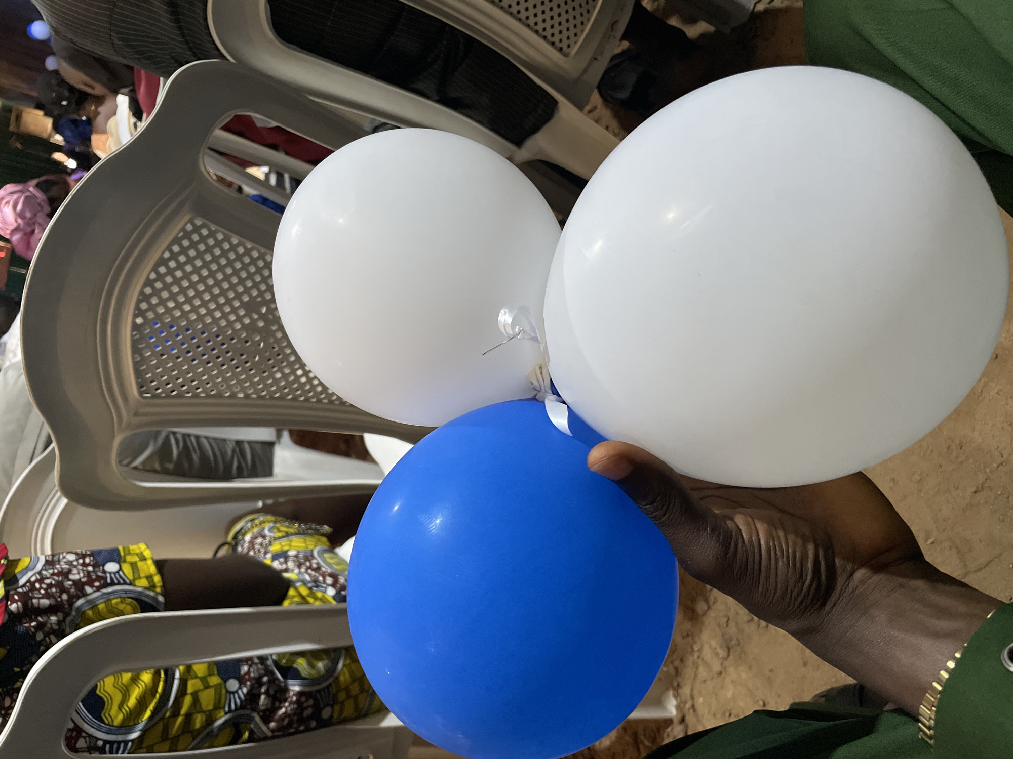 A person's hand, adorned with a gold bracelet, holds three inflated balloons: one blue and two white. The balloons are clustered together, with a small white ribbon visible tying the white balloons. In the background, several white plastic chairs are arranged, some occupied by individuals. One person in the background wears a colorful fabric with a patterned design, possibly traditional attire. The environment appears to be indoors, with a sandy or dirt floor visible. The overall impression is that of a gathering or celebration. The location is specified as Durumi, Nigeria.