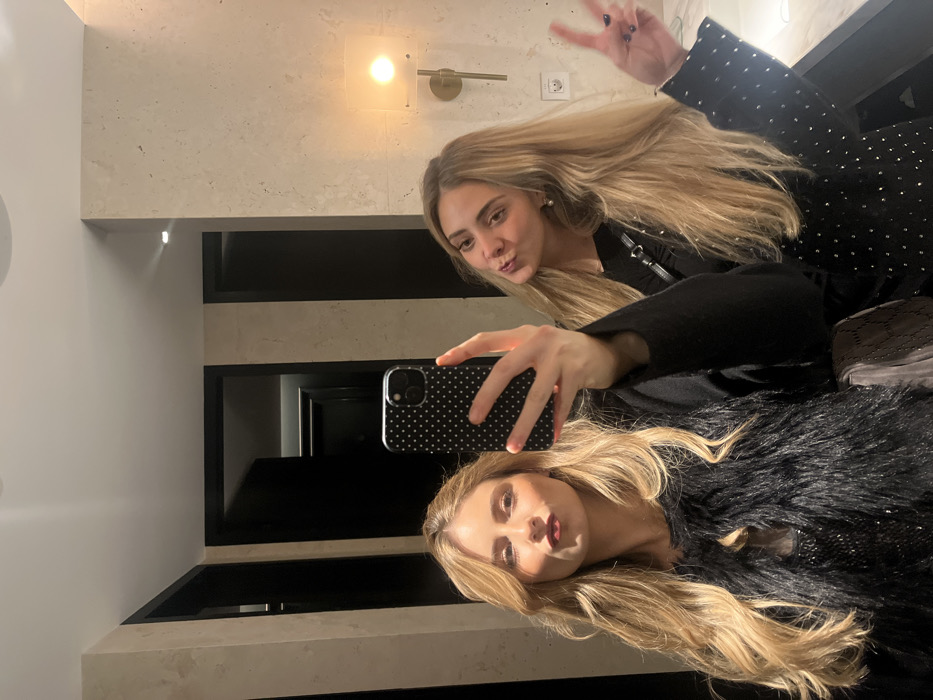 Two blonde women take a selfie in front of a mirrored surface. The mirror reflects an ornate, black and beige wall with recessed shelving. One woman makes a peace sign, and the other puckers her lips. The woman taking the photo wears a fluffy, black vest, while the other wears a black top with silver accents. Both have long, wavy hair. The phone case features a black background with white polka dots.