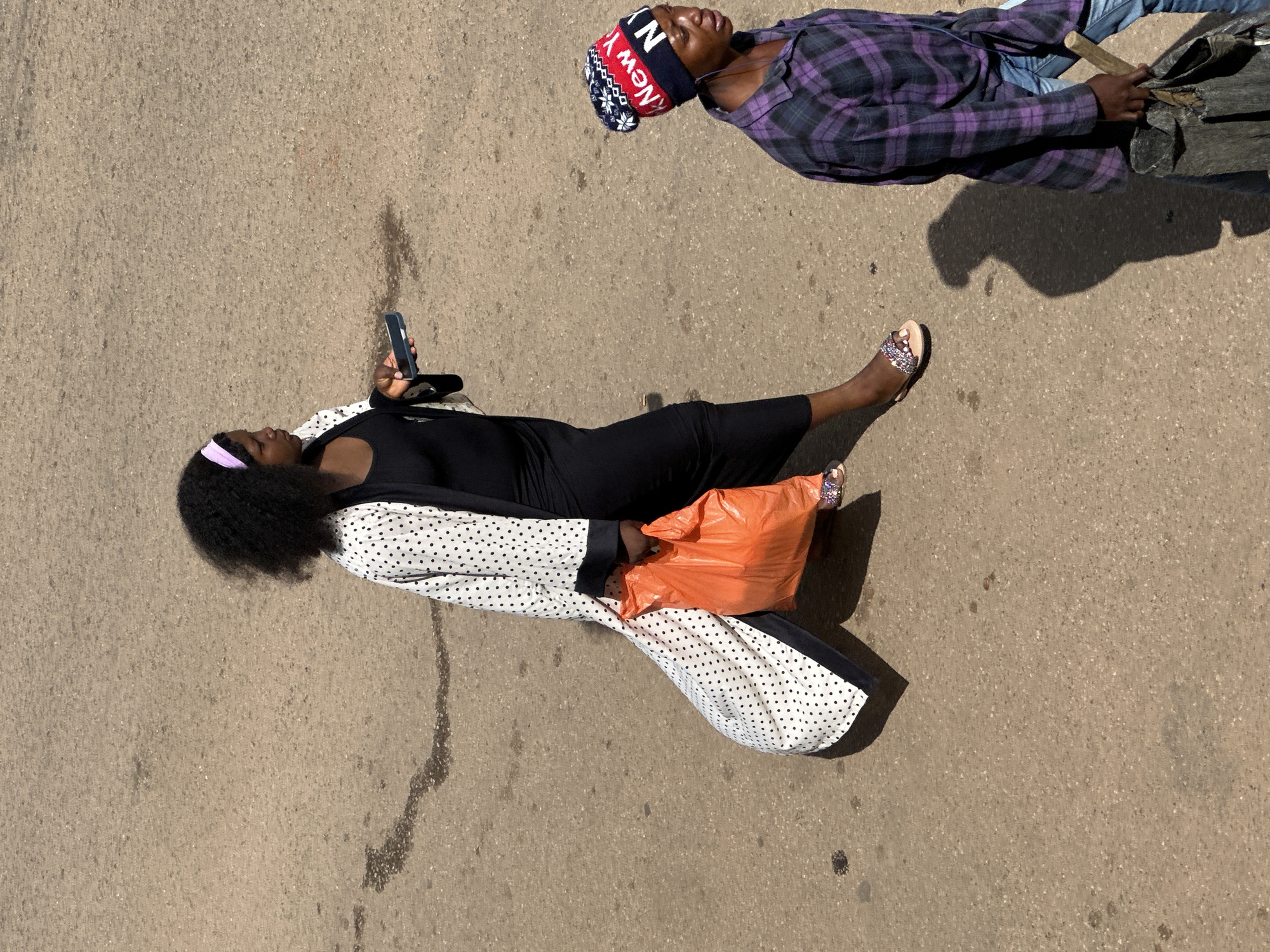 In Bukuru, Nigeria, two individuals are walking on a paved surface. The person on the left is a woman with dark, curly hair, wearing a long, white, polka-dot robe over a black dress. She is holding a bright orange plastic bag in her right hand and a smartphone in her left. She is also wearing sandals. To her right and slightly behind her, another person, identified by a "New York" themed beanie and a purple plaid shirt, is walking. This individual is carrying a bag and what appears to be a broom or stick. The ground surface is a rough, light brown pavement, showing some patches of darker discoloration and a faint shadow cast by the first individual.