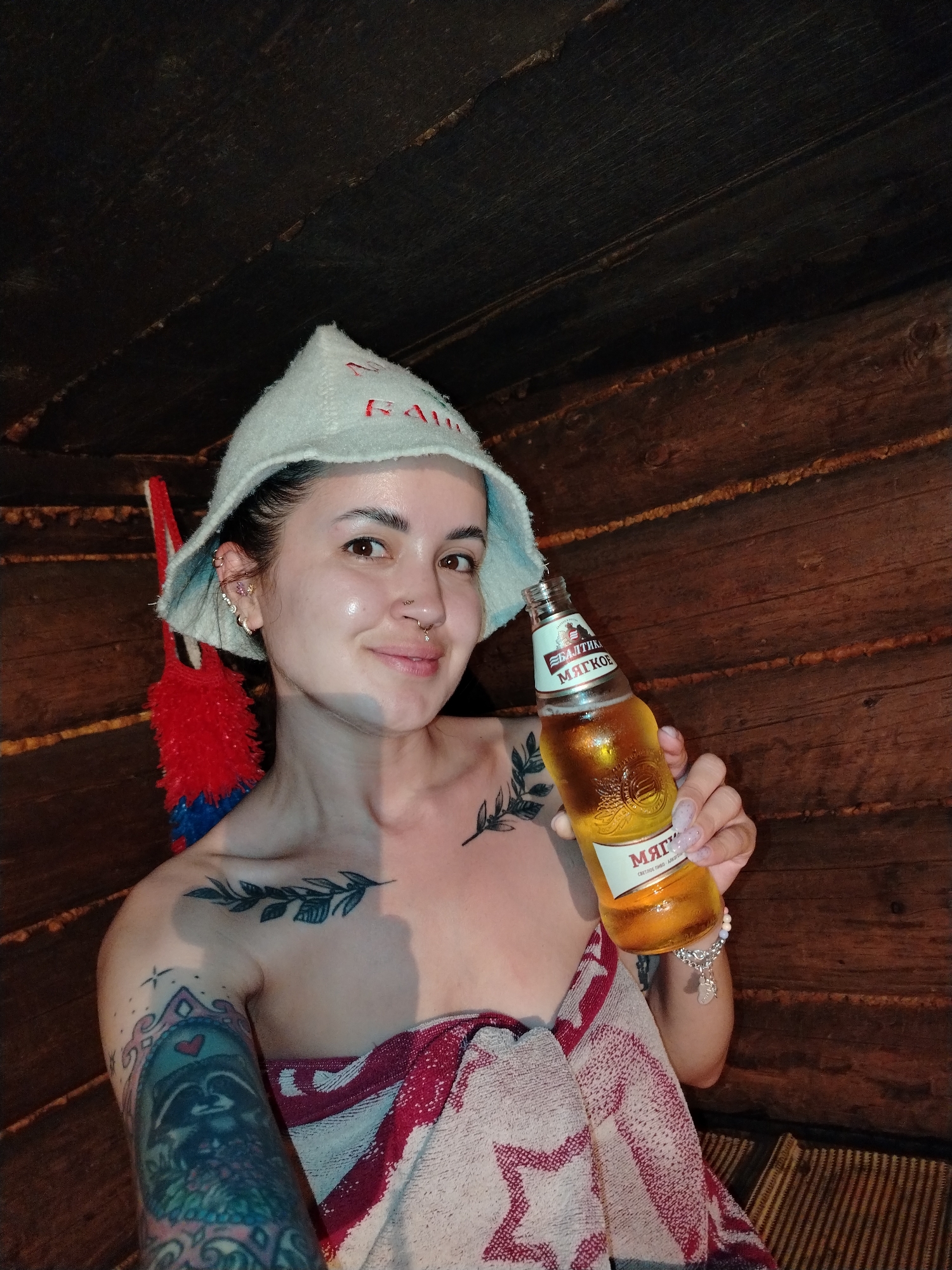 A woman in a sauna holds a bottle of beer, with her body draped in a red and white towel. She wears a white hat with red lettering, and visible tattoos adorn her shoulder and arm. The walls of the sauna are constructed from wooden planks. Based on the labeling on the beer bottle, it is identified as Балтика Мягкое, which is a Russian beer brand. While the precise location in Гожан, Russia, cannot be definitively determined from the image, the sauna environment suggests a relaxed, private setting.