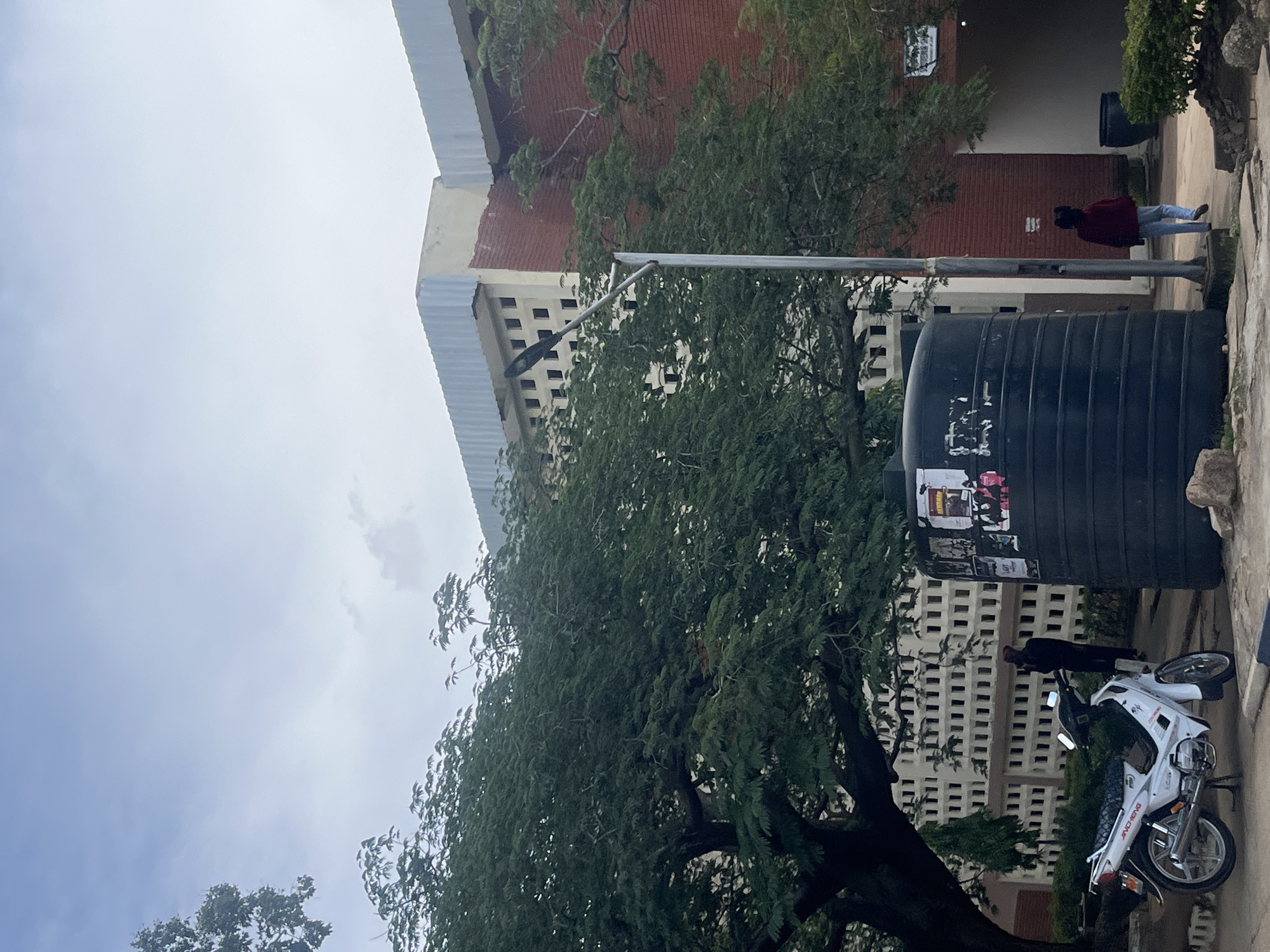 An outdoor, medium shot shows a white motorcycle parked on a paved surface next to a large black water tank. Behind the tank, a person wearing a dark jacket and red head covering stands near the entrance of a building. To the left of the motorcycle, a large tree with dense green foliage provides shade. The background features a building with red brick walls and a white facade with a grid of square openings. A street lamp stands to the right of the tree. The sky is overcast. The scene appears to be on the campus of the University of Jos in Jos, Nigeria.