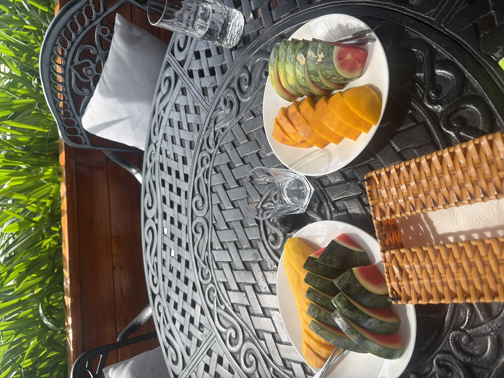 An outdoor table setting at a restaurant in Bắc Nha Trang, Vietnam, features a black, ornate metal table laden with two white plates of sliced fruit, a woven basket, and two clear glasses, one of which is filled with water. The plate in the foreground displays slices of mango and watermelon, while another plate, partially obscured, also holds sliced fruit. Behind the table, a wooden barrier is visible, topped with lush green foliage. The scene is bathed in sunlight, casting shadows on the table and fruit. Two dark metal chairs with light gray cushions are positioned at the table.