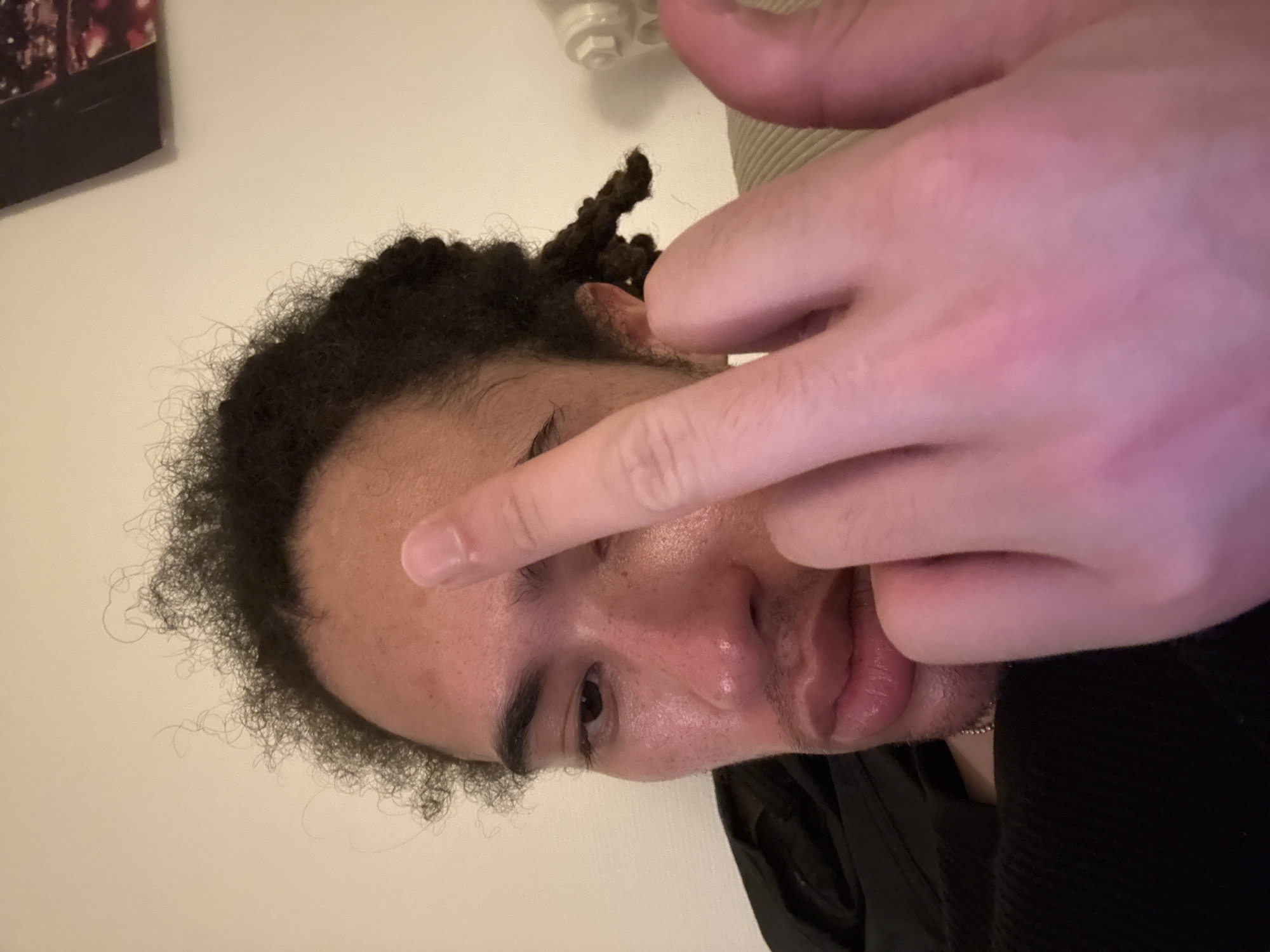 A close-up image depicts an adult male from the chest up in an indoor setting. The individual has a medium skin tone, dark curly hair, with some hair styled into dreadlocks visible on the right side of his head. He exhibits light facial stubble on his chin and upper lip, and a visible mole or blemish on his right cheek. He is wearing a black garment, possibly a sweater or hoodie, and a thin silver chain necklace is visible around his neck. His right hand is raised towards the camera, with the middle finger extended upwards, partially obscuring his forehead and left eye. His visible right eye is open and appears to be looking towards the camera. The background consists of a plain, light-colored wall. A dark, framed object displaying indistinct colorful lights is partially visible in the upper right corner of the frame. To the right of the subject's head, a white, ribbed object, consistent with a radiator, is partially visible. The lighting is soft and appears artificial, casting a slight warmth on the subject's skin. There are no identifiable features within the image to determine the location as Paris, France.