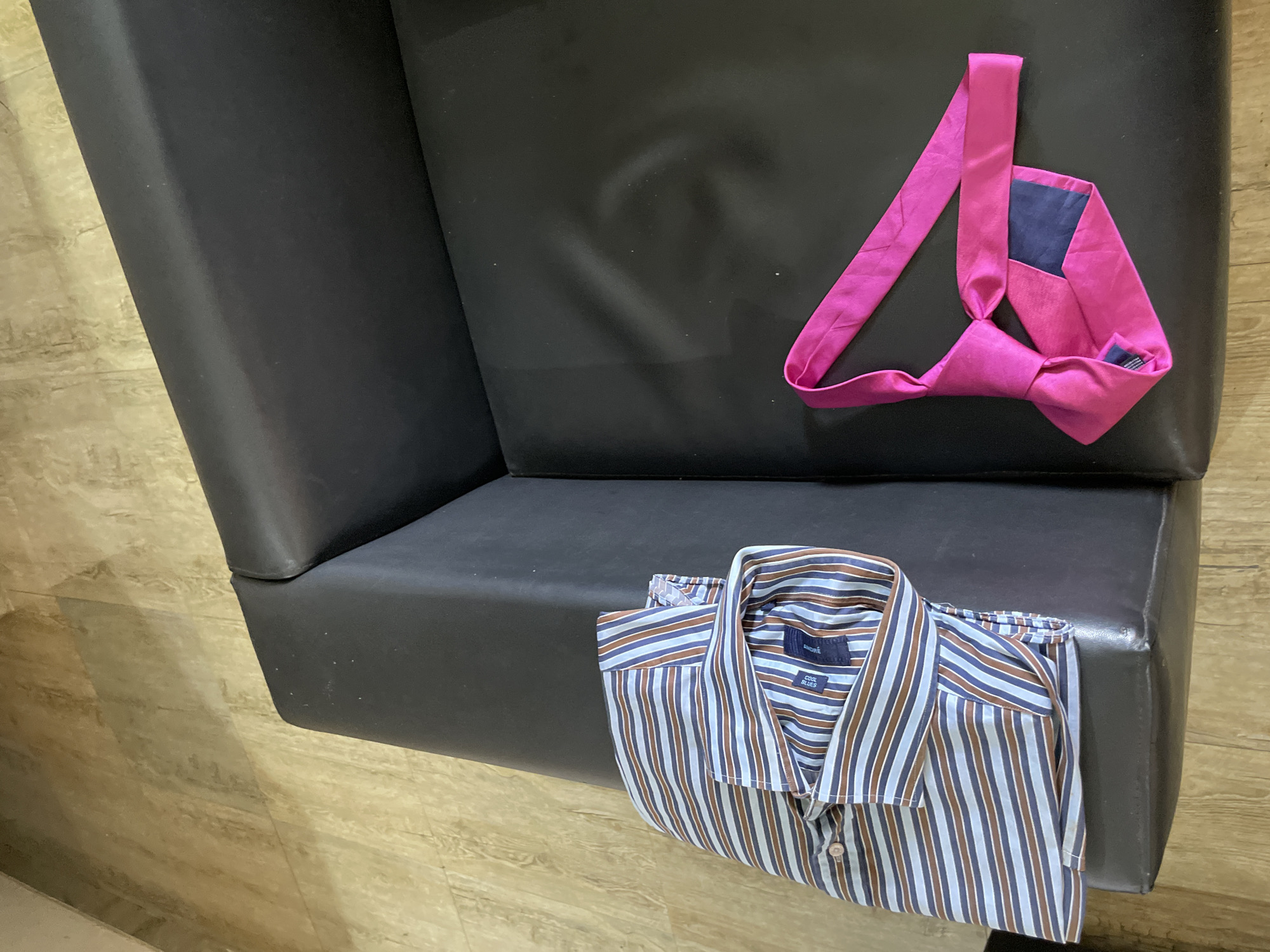A man's striped dress shirt and a bright pink tie are laid out on a black leather couch in a room with wood-patterned flooring. The shirt is folded and placed on the left side of the couch, with its collar and sleeves visible. The tie is knotted and rests on the right side of the couch. The scene appears to be in a residential setting, possibly a bedroom or living room, in Ado, Nigeria.