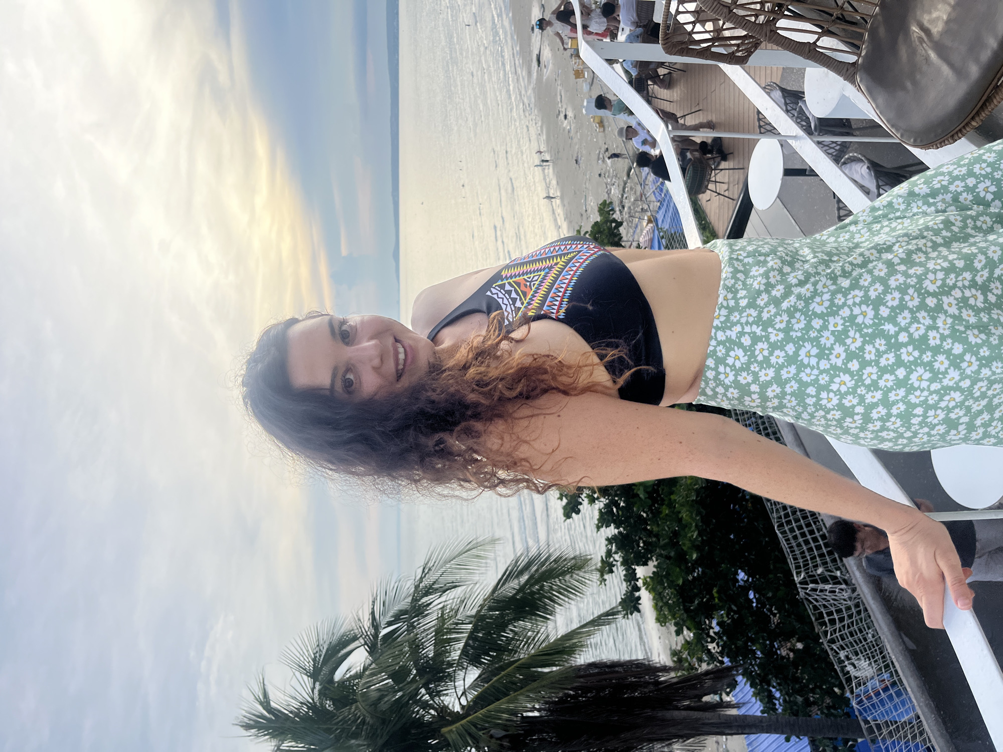 A woman with long, curly brown hair is taking a selfie while leaning on a railing. She is wearing a black bikini top with a colorful geometric pattern and a light green skirt with a white floral print. The woman is smiling at the camera.
Behind her, a beach scene is visible, with calm water and a sandy shore. There are several people in the background, some sitting at tables and others walking on the beach. A palm tree is in the foreground on the left side of the image. The sky is overcast with streaks of light, suggesting it may be early morning or late afternoon. The overall impression is one of a casual, vacation setting.