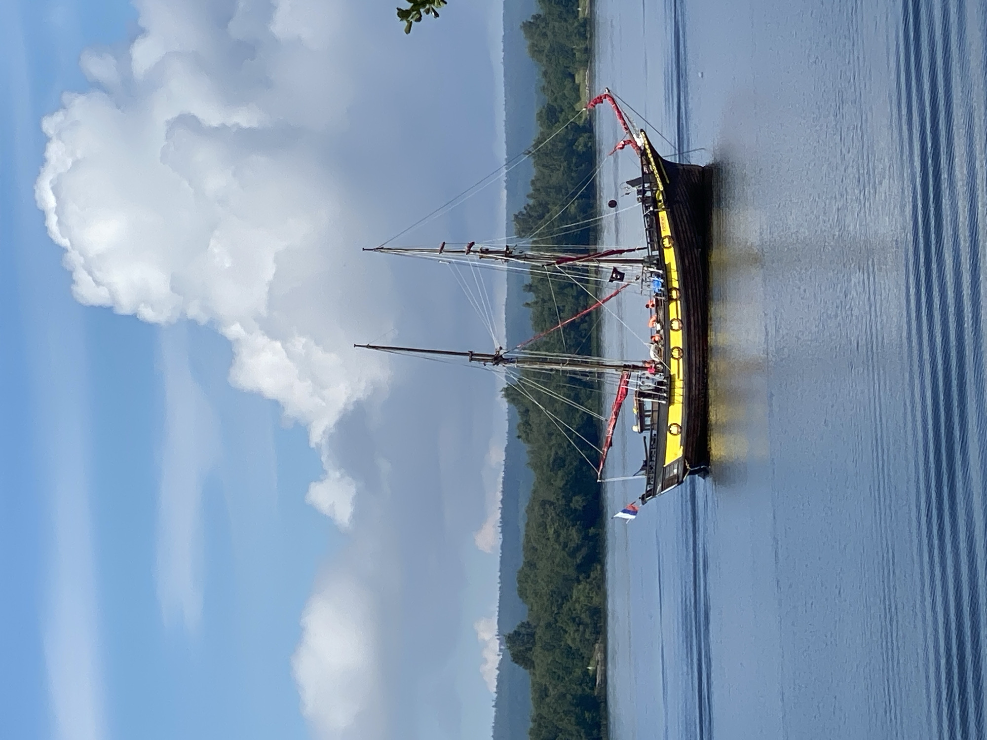 The image depicts a historic sailing ship with two masts, painted yellow and black, sailing on a body of water. The ship has several people on deck and a flag flying from the bow. The background shows a dense forest lining the shore, with a hilly landscape beyond. The sky is blue with scattered white clouds. The image is taken on a clear day, and the water has visible ripples. The scene suggests a calm and pleasant day for sailing, possibly in a natural setting. The ship's design evokes a sense of historical maritime tradition.