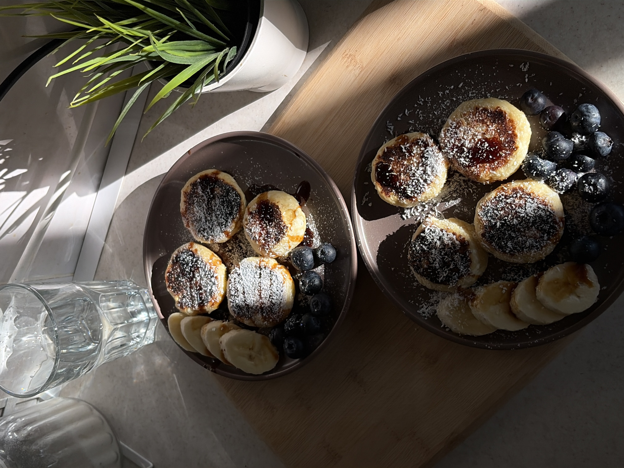 The image shows a top-down view of a breakfast scene, likely indoors. Two plates with what appear to be small pancakes, drizzled with syrup and sprinkled with coconut flakes, are arranged on a wooden cutting board. Each plate also features a serving of blueberries and sliced bananas. To the left of the plate on a countertop, there is a glass with liquid and a potted plant with green foliage is behind. The scene is illuminated by natural light, creating strong shadows. Given the prompt, this scene is likely located in Chaikovsky, Russia. There are no people visible in the image.