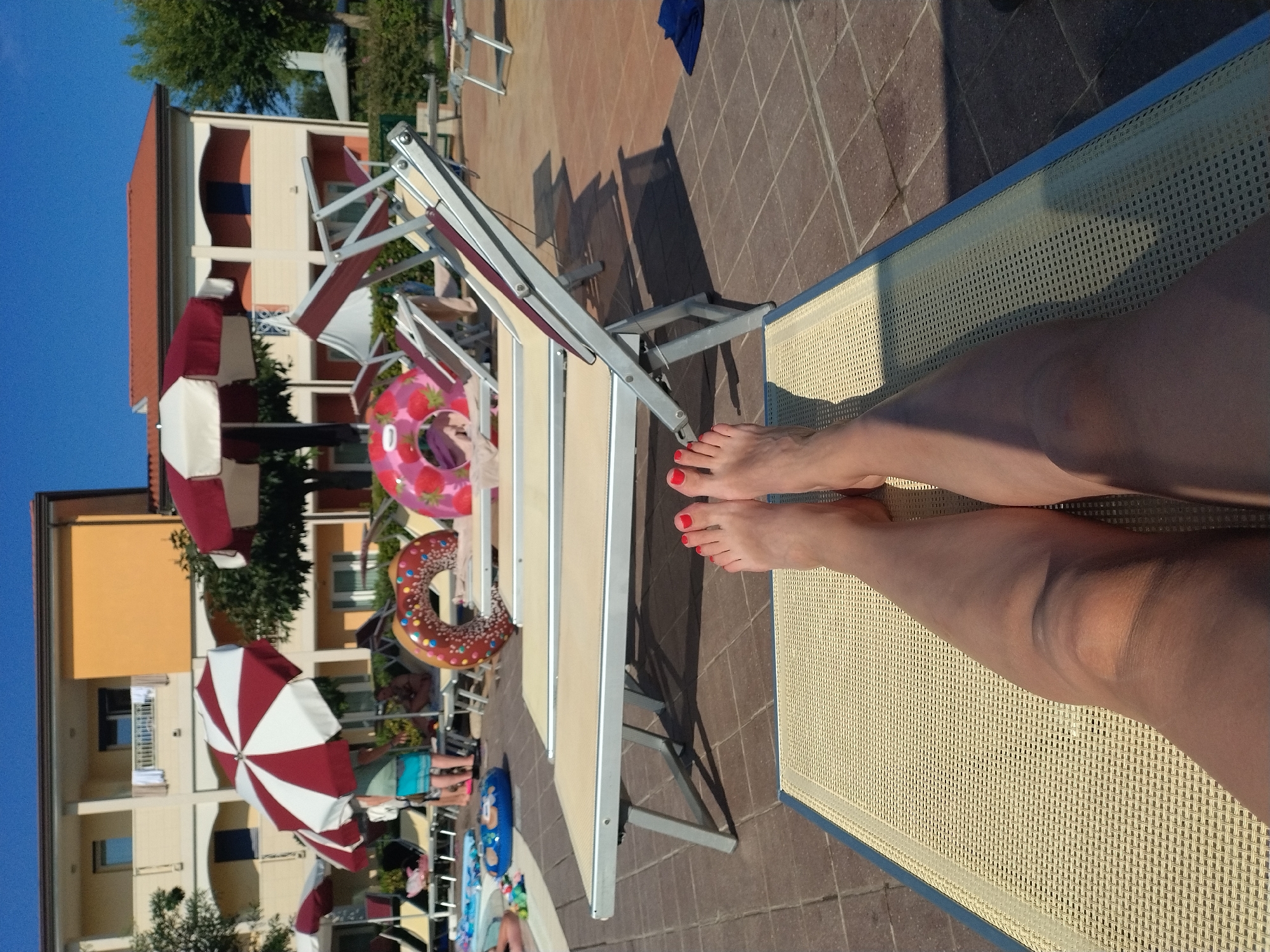 The image depicts a poolside scene in Caorle, Italy, likely at a resort or hotel. In the foreground, a person's legs and feet, adorned with red nail polish, are positioned on a light-colored sun lounger. The lounger has a blue trim and a textured surface. The surrounding area is paved with reddish-brown tiles.
In the middle ground, several other sun loungers are arranged, some occupied by inflatable pool toys, including a donut-shaped float and a strawberry-patterned float. Red and white striped umbrellas provide shade. In the background, a multi-story building with a yellow facade and orange roof is visible, indicative of accommodation. Trees and greenery are also present, suggesting an outdoor setting. The sky is clear and blue, indicating a sunny day.
