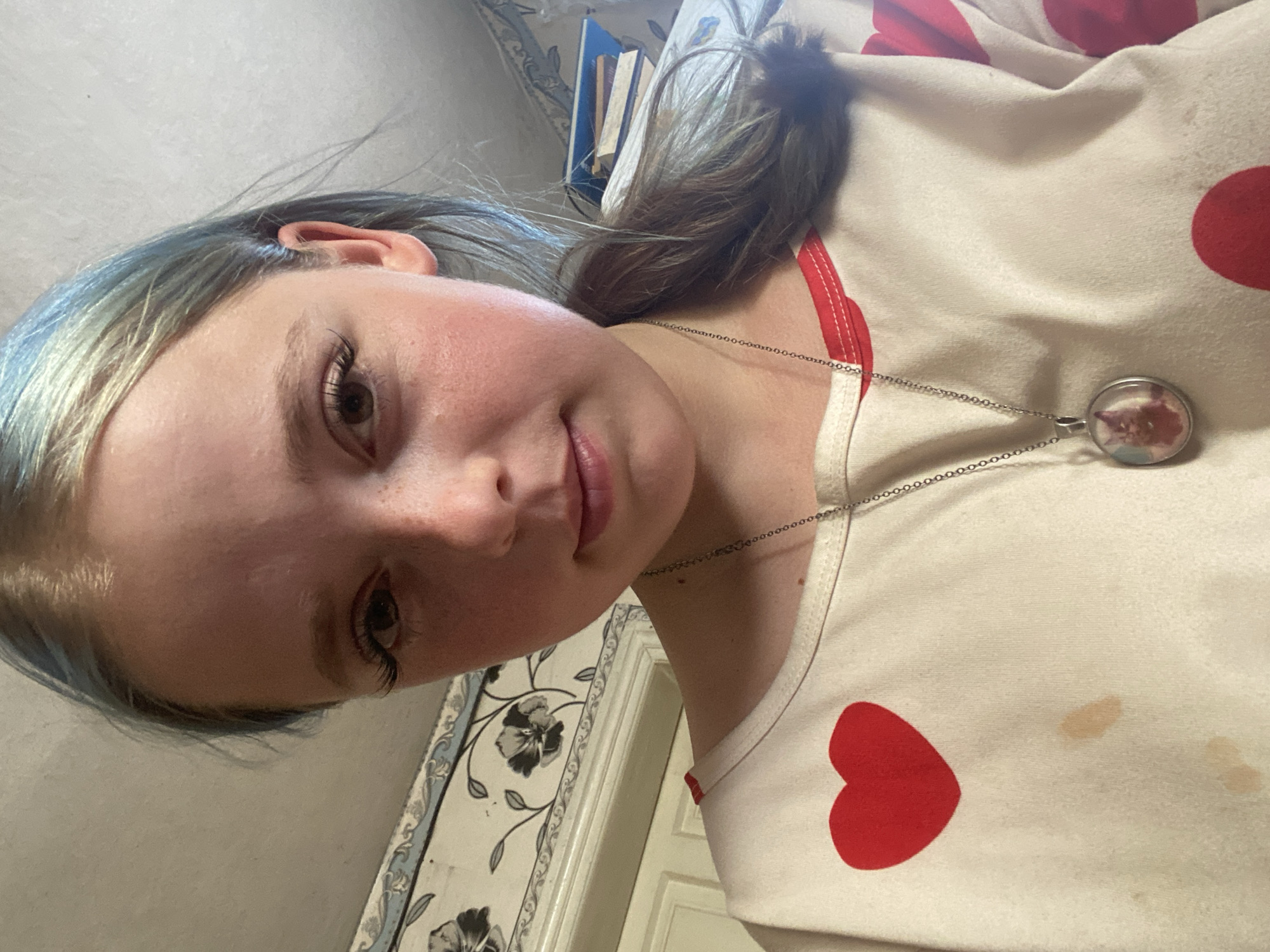 The image is a close-up, slightly tilted selfie of a young person with fair skin and light brown hair that has streaks of blue. They are wearing a cream-colored shirt adorned with red hearts. A silver chain necklace with a circular pendant, which appears to contain a miniature portrait, is visible. The background shows a wall with floral wallpaper and some books on a shelf. The lighting suggests it might be indoors during the daytime. The person's expression is neutral, with a slight smile. There is no visible text in the image. The scene appears to be a casual, personal moment captured in a home environment.