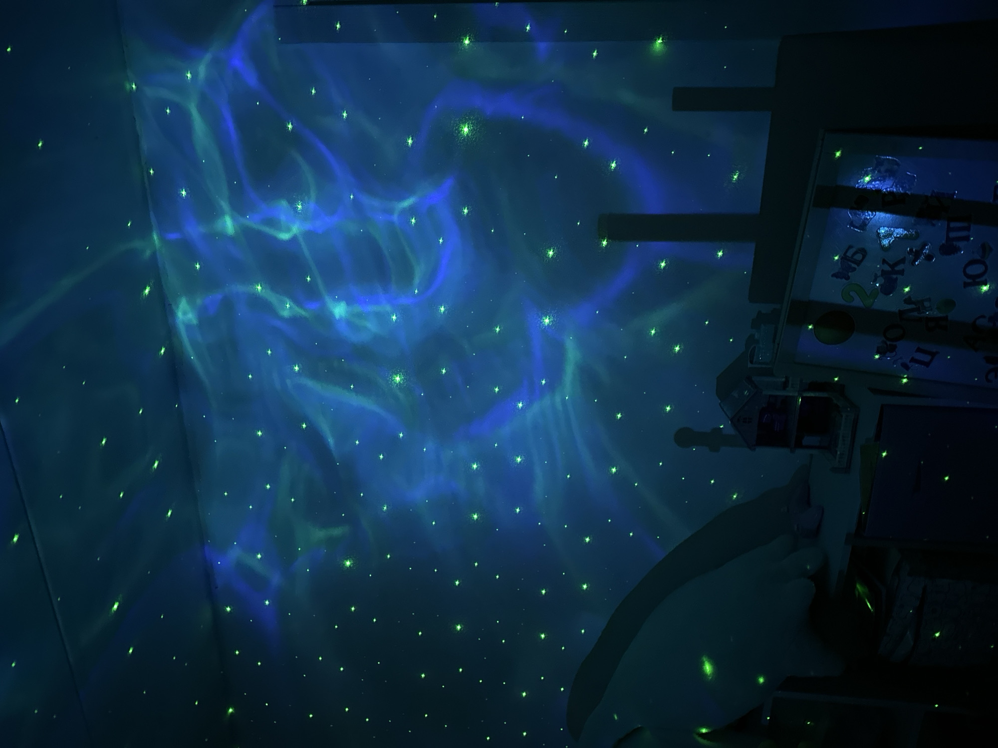 The image shows a dark room, possibly a child's bedroom, in Chaikovsky, Russia, based on the Cyrillic letters visible on a child's learning board. The room is illuminated by a light projector that casts an image of swirling blue and green nebulae and scattered stars onto the wall and ceiling. A portion of a bed or other furniture is visible in the lower right, and a child's learning board or toy hangs on the wall. The scene appears to be indoors and at night.