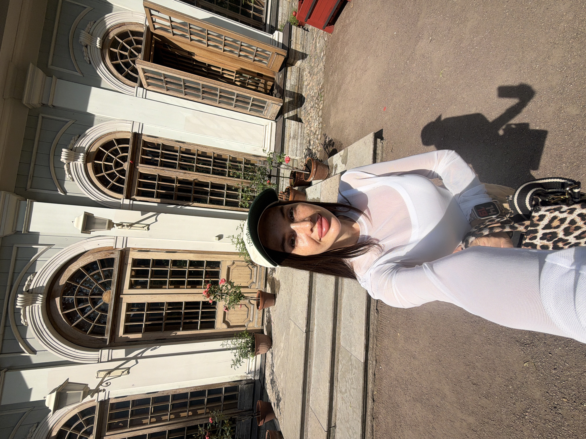 A woman with long dark hair, wearing a white sheer top, tan pants, and a white and green baseball cap, is taking a selfie in front of a building with large arched wooden doors and windows. She is standing on a gravel path, with stone steps leading up to the entrance. Potted plants with pink roses are placed along the steps and in front of the doors. The building has a pale yellow facade with decorative moldings. The image was taken outdoors on a sunny day. Based on the architectural style and the presence of the building's unique design, this scene is likely located at the Tauride Garden in Saint Petersburg, Russia.