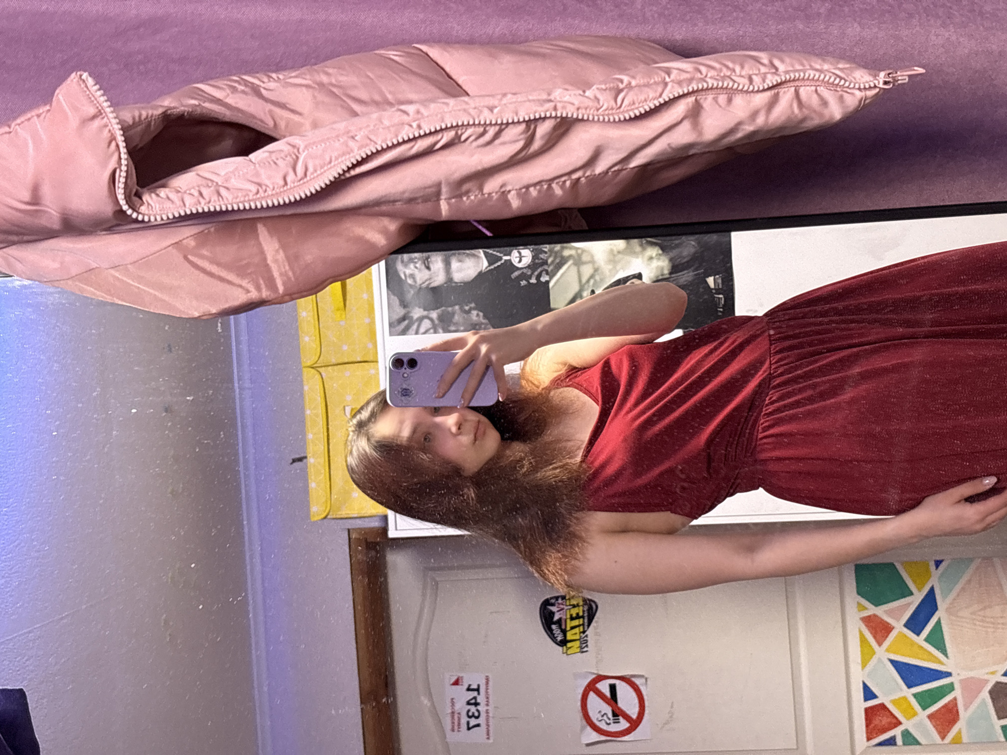 A mirrored selfie of a young woman in a red dress, indoors. She is holding a purple phone, presumably taking the picture. The background includes a pink jacket, a poster on the mirror, a yellow item, a door with a "no smoking" sign, and a colorful geometric artwork. The location is implied to be Прикамский, Russia, although this is not directly evident from the image.