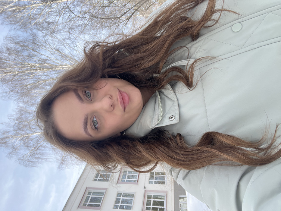 A selfie-style photograph captures a young adult female outdoors in Chaikovsky, Russia, during winter. The subject, positioned centrally, has long, wavy brown hair, light-colored eyes, and fair skin. She is wearing a light mint green puffer jacket with a fastened collar and has a silver hoop earring visible on her left ear. Her expression is a slight smile with subtly pursed lips, looking directly at the camera. In the background to the left, a light-colored, multi-story building with numerous rectangular windows is partially visible. The ground in front of the building is covered with a layer of snow. To the right and extending above the subject, several bare deciduous trees with slender, light-colored trunks are visible against a pale blue sky. The overall environment indicates a cold, snowy outdoor setting.