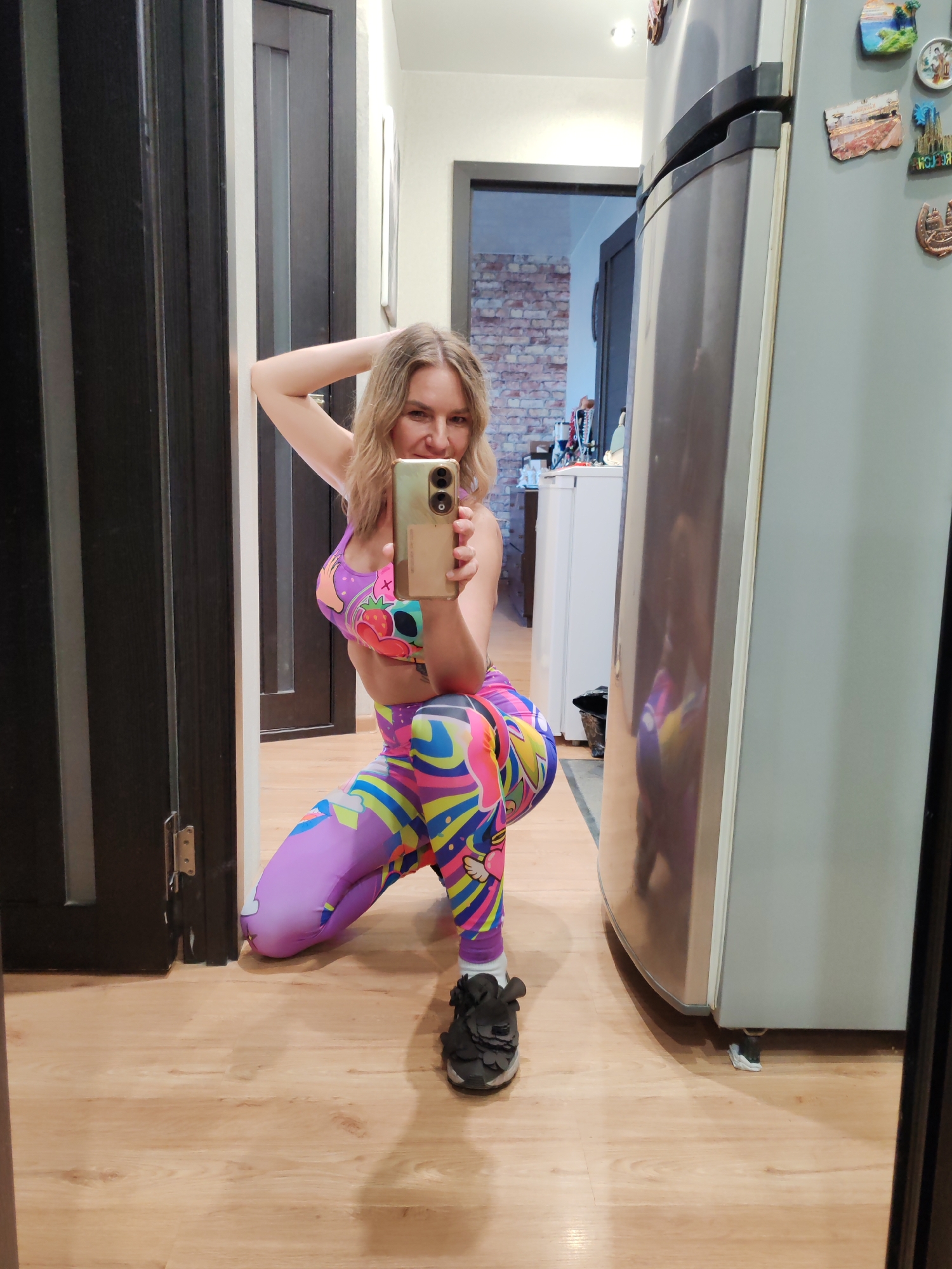 A woman is taking a selfie in what appears to be a home interior, likely a kitchen. She is kneeling, facing the camera, with her right hand raised behind her head and her left hand holding her phone. She is wearing a colorful, patterned athletic outfit consisting of a sports bra and leggings, along with black sneakers.
Behind her, there's a doorframe and another room visible with a brick wall and white furniture. To her right is a refrigerator with magnets on it. The floor is made of light-colored wood.
There is no information available in the image that indicates the location to be Petropavlovsk-Kamchatsky, Russia.