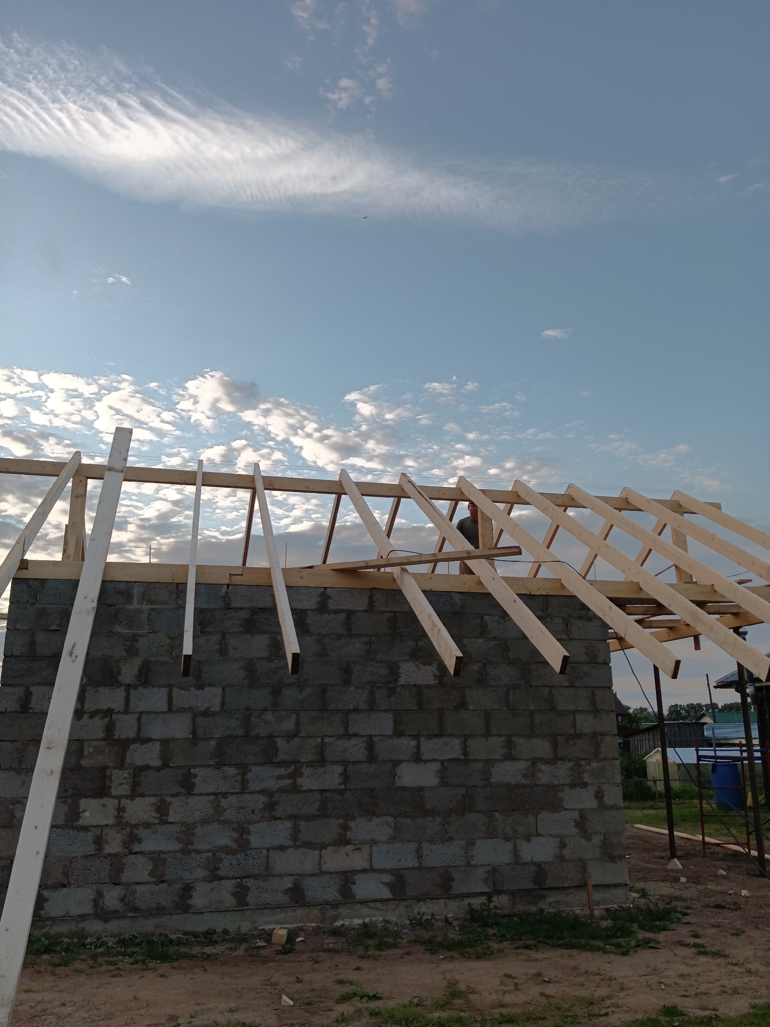 The image depicts the construction of a roof frame on a cinder block building in the city of Foky, Russia. A person is visible near the center of the image, standing on the roof structure. The sky above is partly cloudy with wispy, elongated clouds. The foreground shows dirt and grass. The overall scene suggests an ongoing construction project.