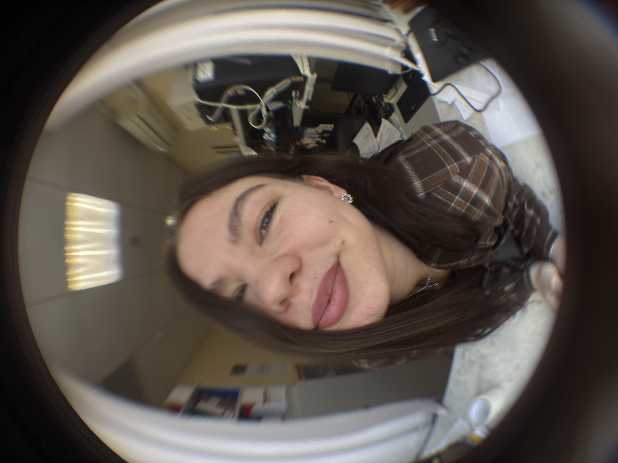 The image is a fisheye-lens selfie of a fair-skinned young woman with brown hair, wearing a brown plaid shirt. She is smiling slightly at the camera. The background appears to be an office environment. Desks with computers and paperwork are visible. A window lets in natural light. An air conditioning unit is mounted on the wall. Based on the location details, the scene is in Perm, Russia.