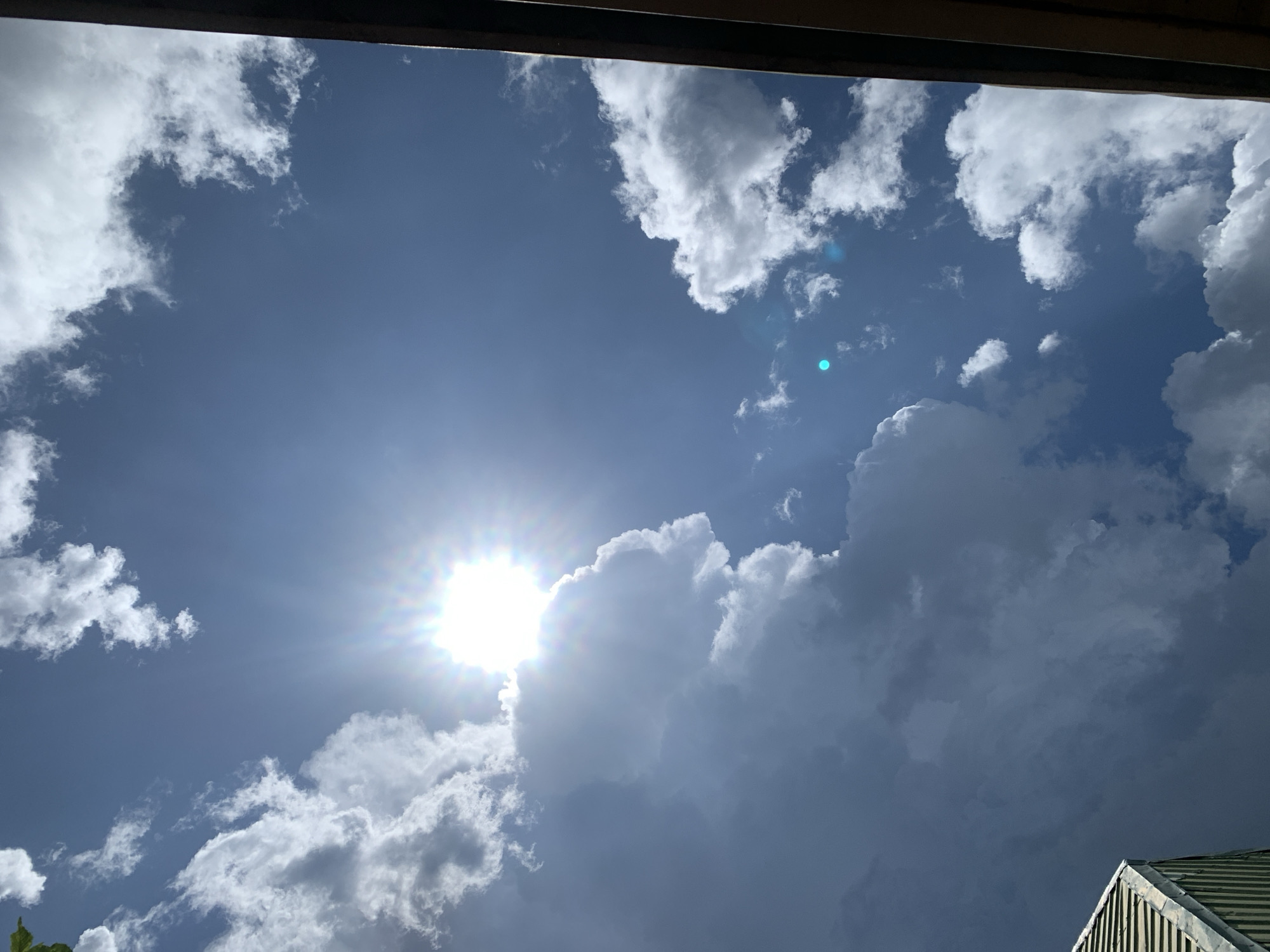The image depicts a bright, sunny sky with scattered clouds, viewed from a low angle. The sun is visible, casting a strong glare and creating lens flare effects. The sky is a deep blue, with fluffy white and gray clouds dominating the frame. A portion of a building's rooftop, with green corrugated metal, is visible in the lower right corner. The scene appears to be an outdoor setting, likely in Kuru Karama, Nigeria, given the context. There are no people or animals visible in the image.