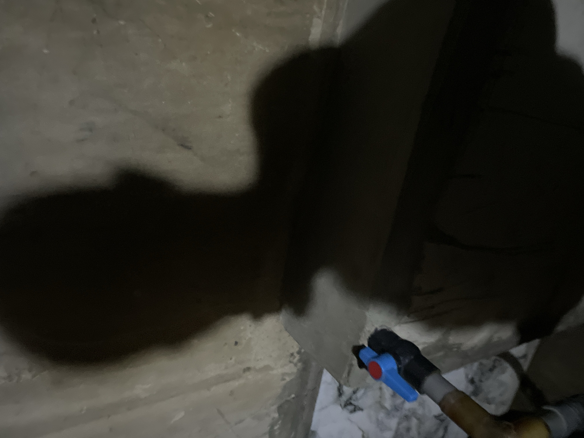 The image shows a dimly lit corner of an interior space. The walls are textured and appear to be made of concrete or plaster. A pipe with a blue and red handle is visible, running along the base of the wall. Marble floor tiles are below the pipe. The lighting is poor, casting large shadows on the walls and obscuring details.