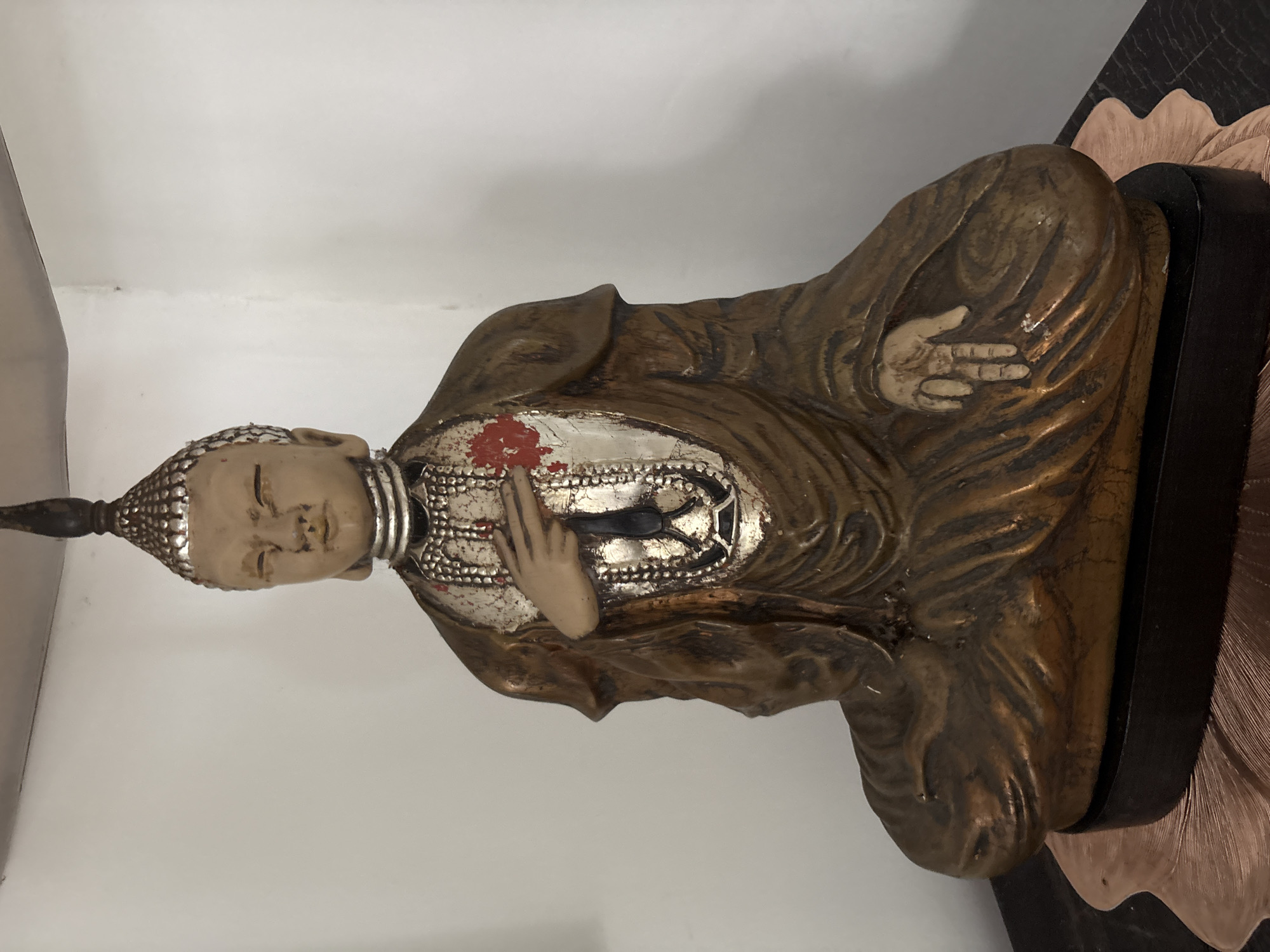A close-up, front-facing medium shot captures a weathered, seated Buddha statue positioned in a corner of a room. The statue, rendered in a gold and tan color palette with hints of red and silver embellishments, is seated in a lotus position. Its right hand rests on its knee, palm facing upwards, while its left hand is raised to its chest, fingers splayed. A decorative, ornate chest piece with layered beadwork and a red accent adorns the statue. The statue is situated on a dark wooden surface, possibly a table, which is layered with a decorative, fan-shaped mat. The background consists of a plain, off-white wall, with the corner of the room visible. The lighting appears to be artificial and soft, casting subtle shadows. The scene does not depict any specific location within Nha Trang Ward, Vietnam, beyond being an indoor setting.