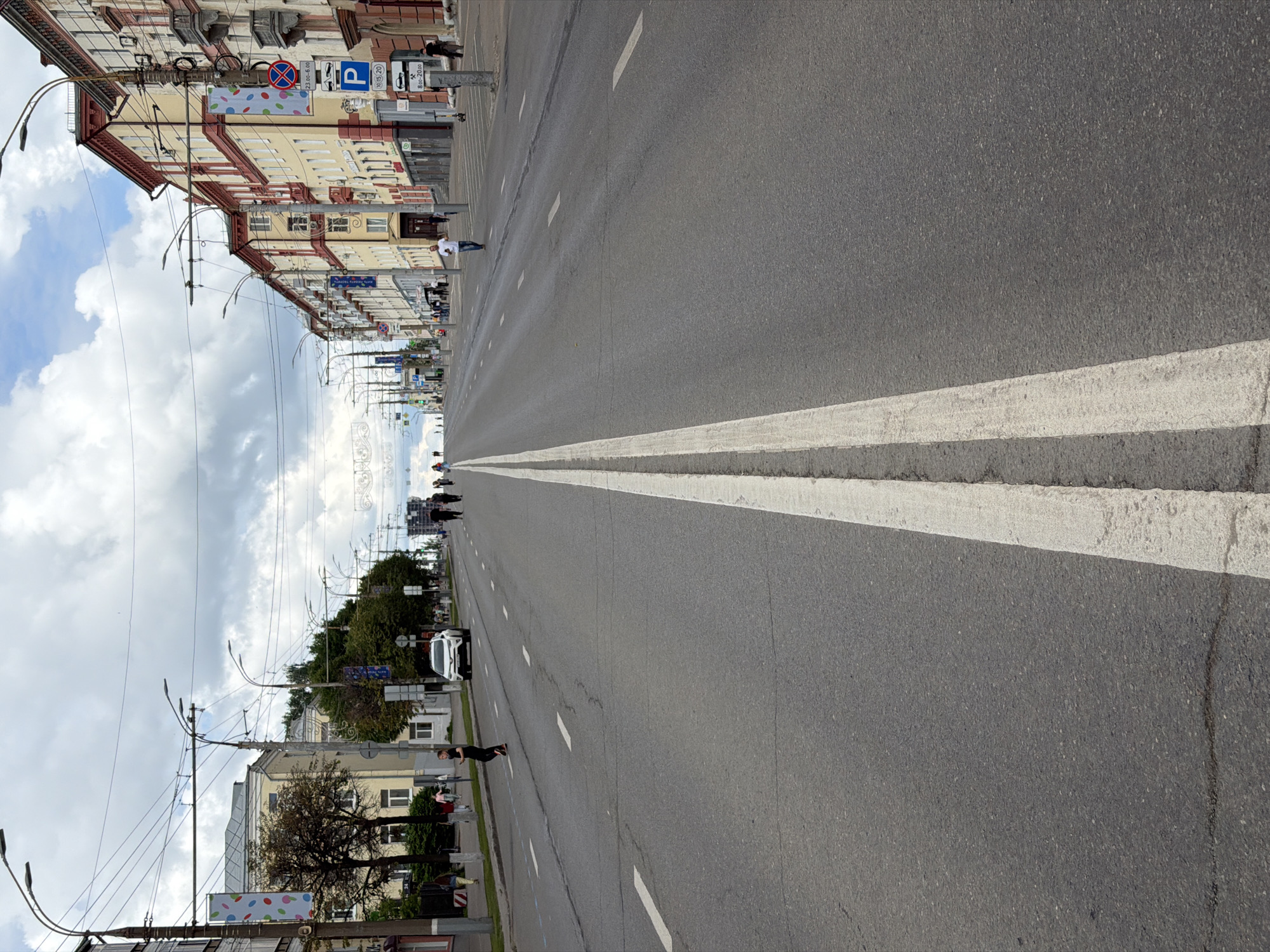 The image shows a wide, multi-lane paved road in Izhevsk, Russia, likely a city street, with buildings lining the right side and trees and a sidewalk on the left. A prominent double white line divides the lanes in the foreground. Several people are visible walking on the sidewalk and crossing the street, and a white car is parked on the left side. Overhead, electrical wires and streetlights crisscross the sky, which is partly cloudy. The architecture of the buildings suggests an urban setting, with decorative balconies and varied window designs. Street signs, including a parking sign with times, are visible on the right.