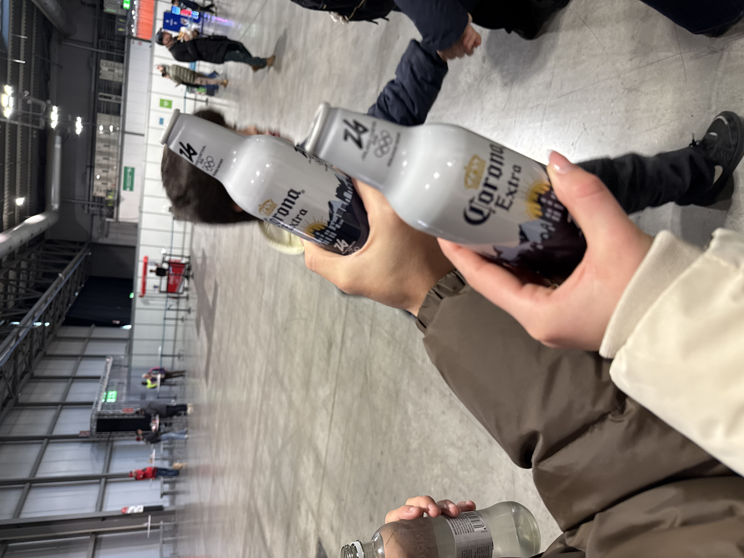 The image depicts an indoor scene within a large exhibition hall, characteristic of Fiera Milano in Rho, Italy. In the immediate foreground, two hands are prominently displayed, one holding two bottles of Corona Extra beer and the other holding a clear glass bottle of water. The Corona bottles are white and feature branding including "Corona Extra," a stylized skyline, and "Milano Cortina 2026" with the Olympic rings, indicating a special edition related to the upcoming Winter Olympics. The hand holding the Corona bottles is partially obscured by a child's head and shoulder on the right, clad in a dark blue jacket. The hand holding the water bottle is on the left, connected to an arm wearing a brown jacket.
The background shows a vast, open space with a light gray concrete floor and high ceilings supported by exposed metal beams and industrial lighting fixtures. Numerous individuals are visible in the mid-ground and background, appearing blurred. They are dressed in various casual attire, including jackets and pants. Some individuals are standing or walking, while others are gathered around small, light-colored tables set up with temporary barriers. One person in a red jacket stands near a large vertical panel. Distant booths and structures with illuminated signs, some displaying green and red lights, are also present, suggesting an event or exhibition. The overall environment is brightly lit.