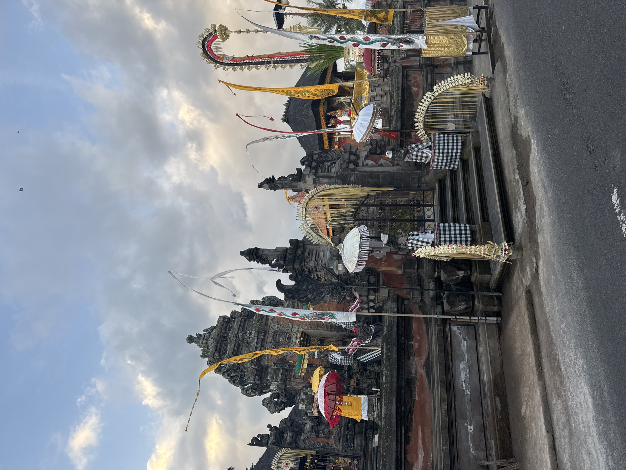 The image shows a temple in the city of Blahbatuh, Indonesia, adorned with festive decorations. The architecture is traditional Balinese, featuring stone carvings and tiered roofs. Numerous flags in yellow, white, and red are displayed, along with ornate umbrellas, some red and white, others white. Two figures in traditional attire, possibly statues or effigies, are visible flanking the entrance, dressed in black and white checkered cloths. Archways decorated with floral motifs and golden fabric frame the entrance. The ground in front of the temple is a paved street, showing signs of wear and tear. The sky above is partly cloudy, with the sun casting a soft, warm light, suggesting it is either early morning or late afternoon. Two small, dark objects, possibly kites, are visible in the distant sky. The overall impression is one of a culturally rich and visually vibrant setting, likely during a festival or celebration.