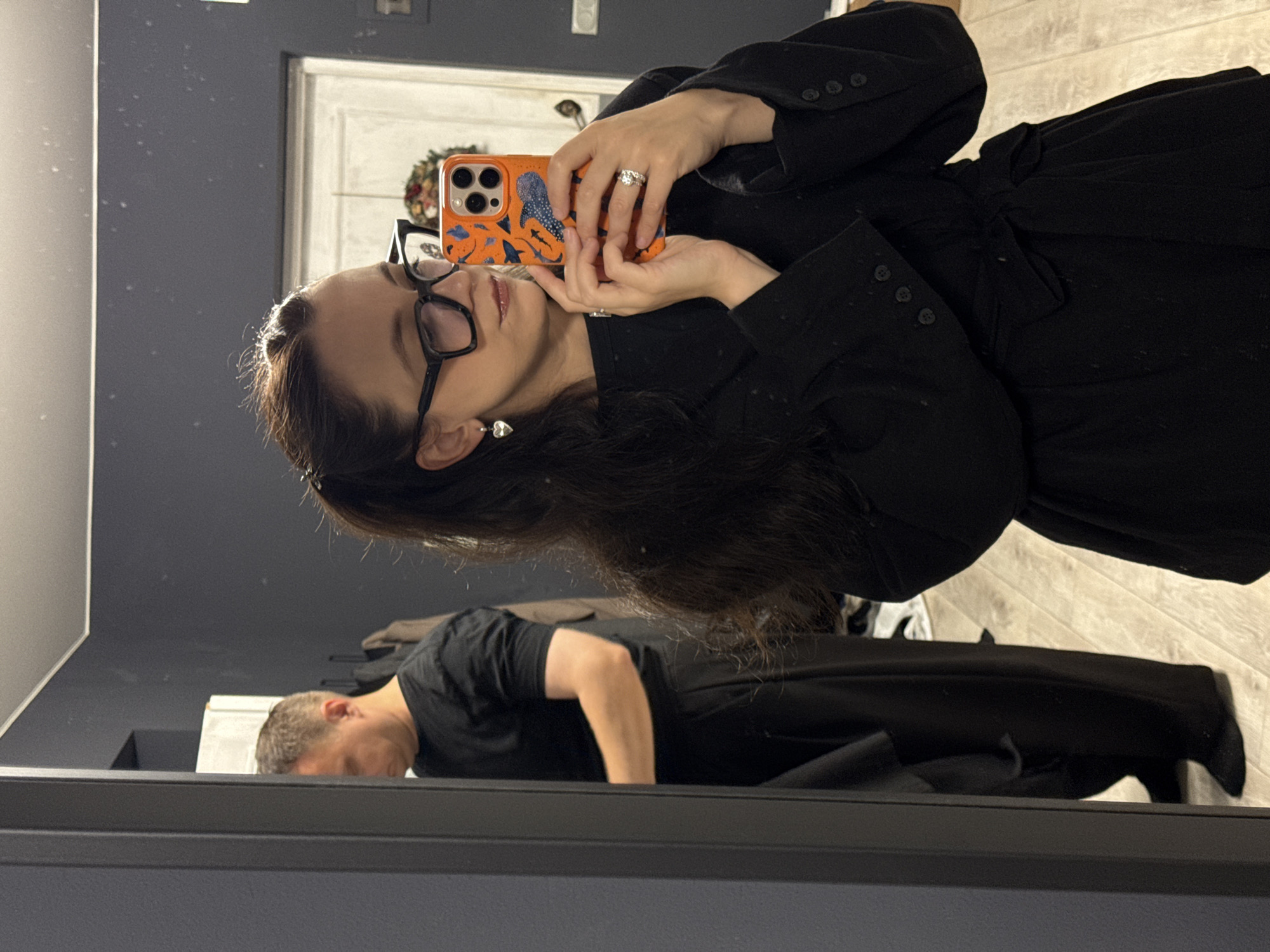 A woman is taking a mirror selfie in an indoor setting, likely a home. She is wearing a black outfit and glasses, and holding an orange phone with a shark-themed case. A man in a black t-shirt and pants is reflected in the mirror behind her. The environment appears to be a room with dark walls and a light-colored wooden floor. There is a doorway and some clothing hanging in the background. No specific location within Chaikovsky, Russia, can be identified from the image.