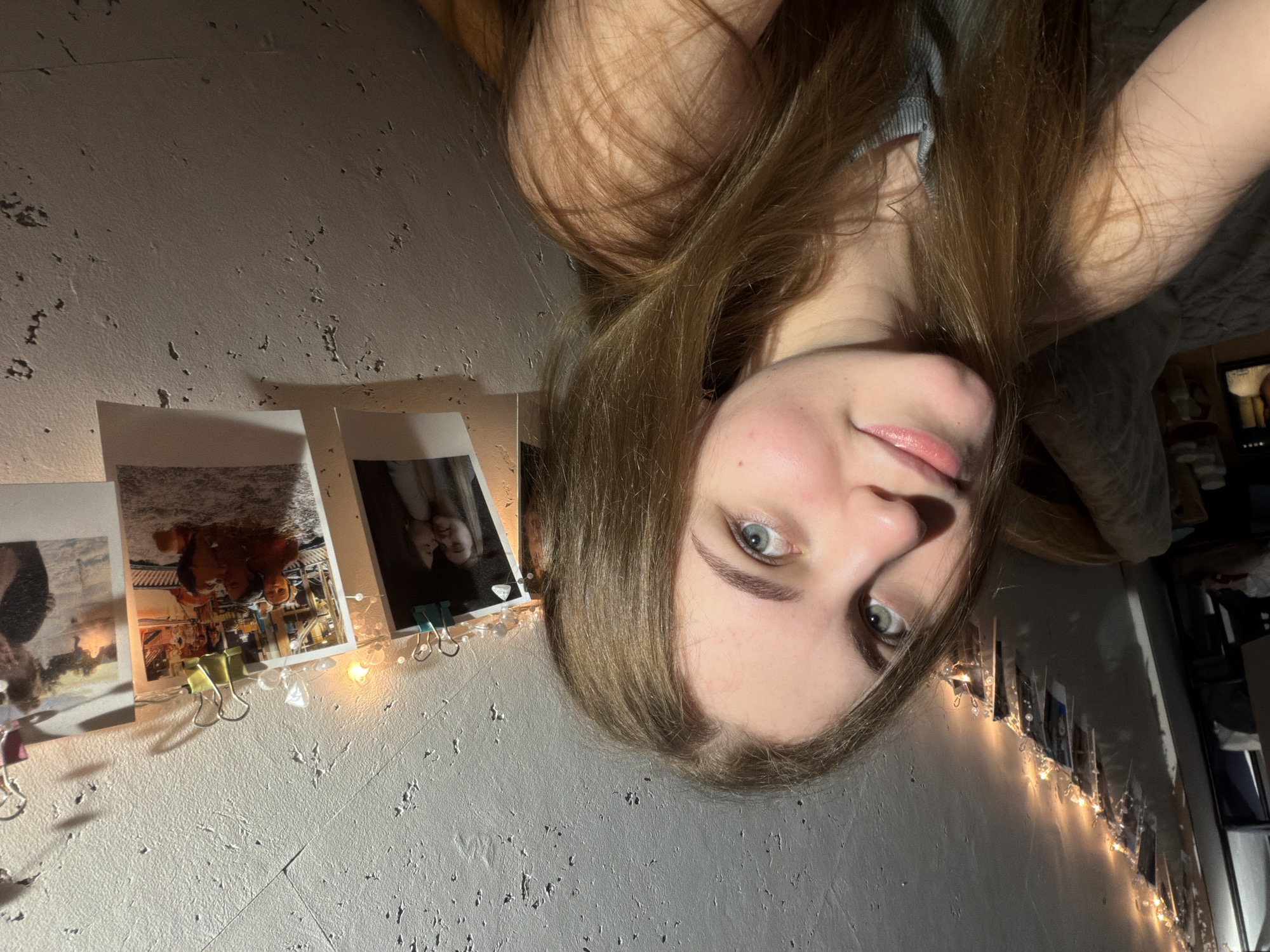 The image shows a young woman with long, light brown hair and blue eyes, taking a selfie in what appears to be a bedroom. She is leaning towards the camera, with her left shoulder visible. The background consists of a textured wall decorated with string lights and several printed photographs clipped to the wall with binder clips. Some of the photographs appear to show people, potentially friends or family. The lighting in the room is dim, with the string lights casting a warm glow. There are no specific indicators in the image to suggest a location in the city of Dnipro, Ukraine.