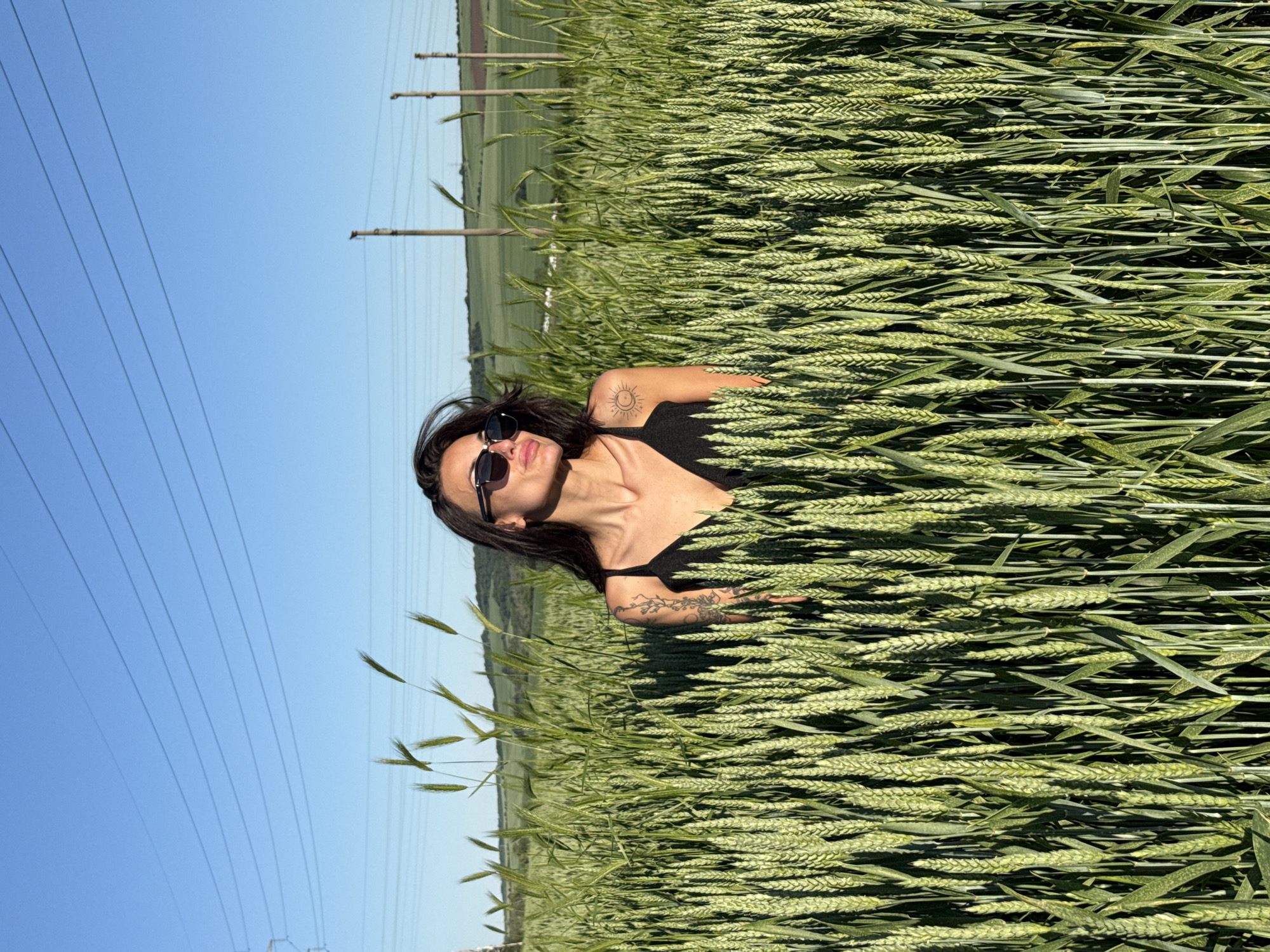 A woman lies in a field of green wheat under a clear blue sky. She wears sunglasses and a black top and has tattoos on her arms and shoulder. Overhead power lines are visible in the distance, indicating an agricultural area. The photo was taken in a location in the city of Pleven, Bulgaria.