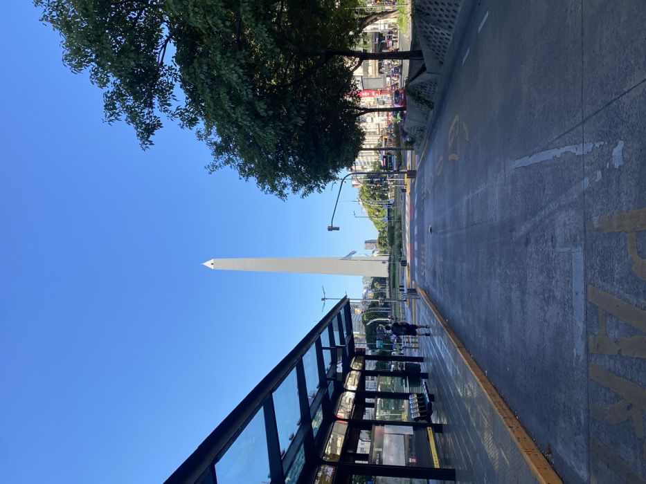 A tall, white obelisk stands prominently in a bright, blue sky. A tree canopy overhangs the scene. A bus stop with glass panels and metal framing is visible in the foreground. The street appears freshly cleaned, reflecting sunlight. Distant buildings and traffic signals line the thoroughfare leading towards the obelisk. Two figures are standing near the bus stop, seemingly waiting.