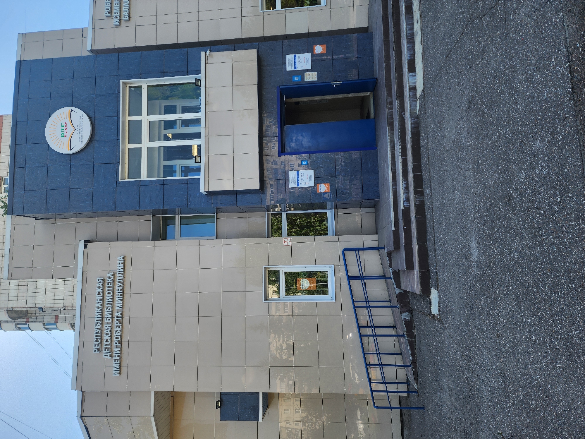 The image depicts the exterior of the Republican Children's Library named after Robert Minnullin in Kazan, Russia. The building has a facade with light beige and blue-gray tiled sections. A circular sign with a colorful emblem is affixed to the blue-gray portion of the building. An open blue door is also visible on the blue-gray part. The library’s name is vertically displayed on the light beige section. A blue handrail suggests the presence of steps leading to the library. The street is made of asphalt. The sky is blue.