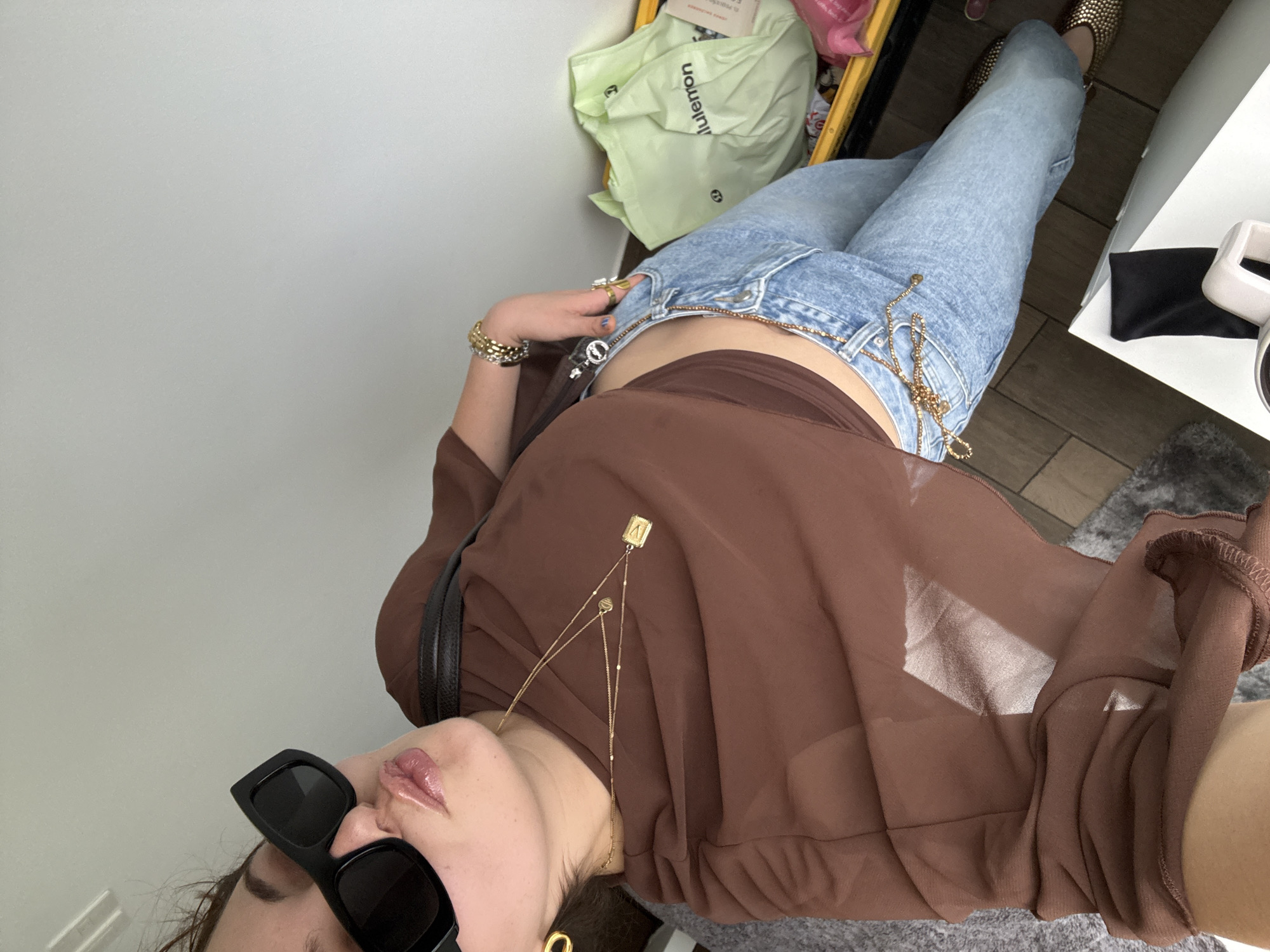 A woman poses for a photograph in what appears to be an indoor setting. She is wearing jeans, a brown top, and sunglasses, and jewelry including gold-toned necklaces and a bracelet. A green bag with the word "lululemon" on it is visible in the background, as well as a few other obscured items in a yellow bin. The woman's face is angled towards the camera, while her body is positioned with her legs stretching away from the camera in the direction of what appears to be a white table, a small black bag, and tiled flooring. The setting includes gray carpeting near the camera's location and white walls. The location is not discernible based on the provided image.