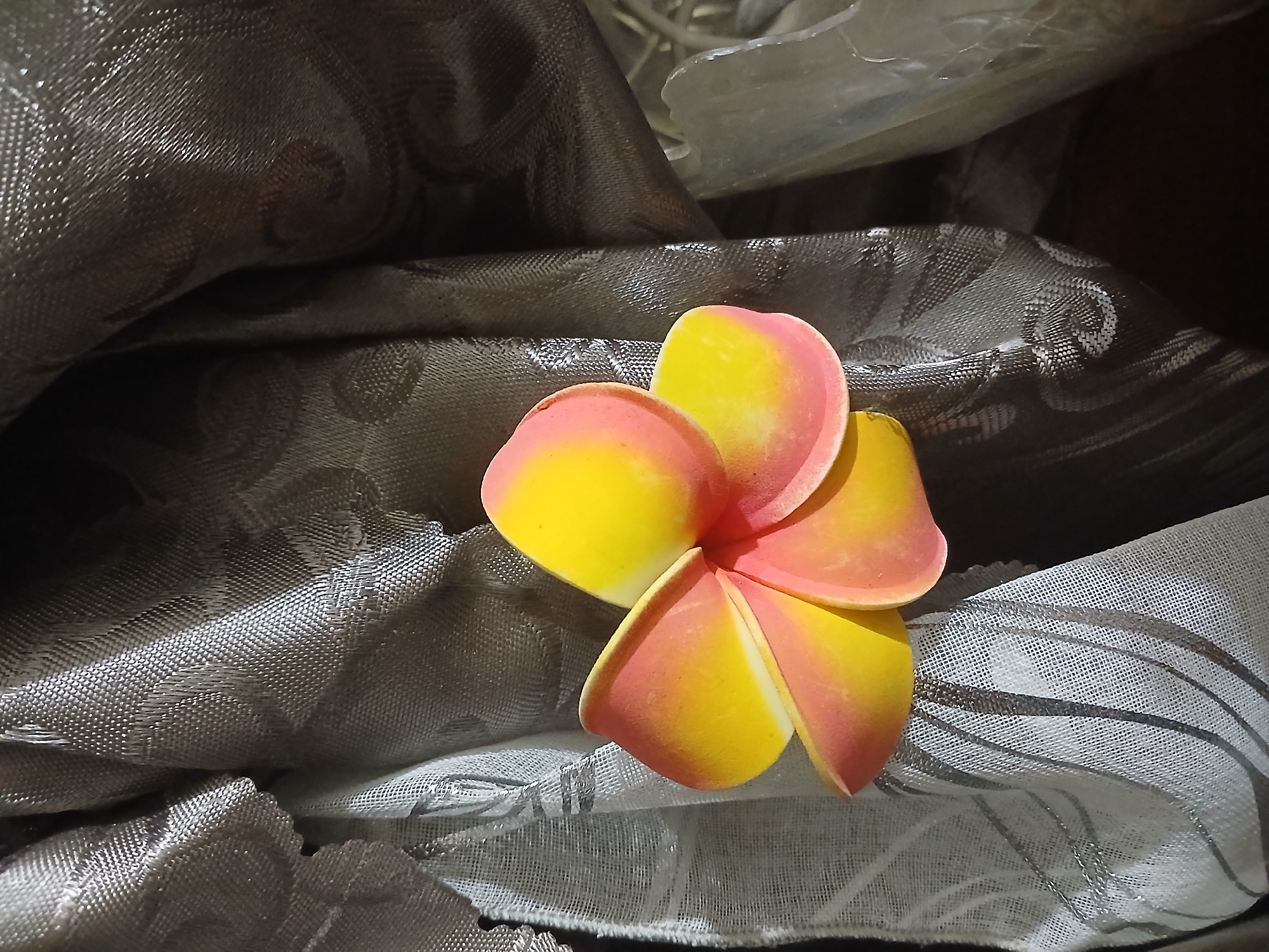 A close-up, eye-level shot focuses on a single, artificial plumeria flower with yellow and pink petals resting on a textured, dark grey fabric. The fabric is draped and folded, creating soft shadows and highlights that emphasize its woven pattern. In the background, other draped fabrics and some blurry, translucent materials are visible, suggesting an indoor setting. The lighting appears to be artificial, casting shadows and illuminating the flower and fabric with a warm glow. The composition is centered on the flower, with the surrounding fabric providing a contrasting texture and color. No people, text, or specific location cues are visible within the frame, beyond the provided context of Ijevan, Armenia. The scene is static, with no discernible activity or event taking place.