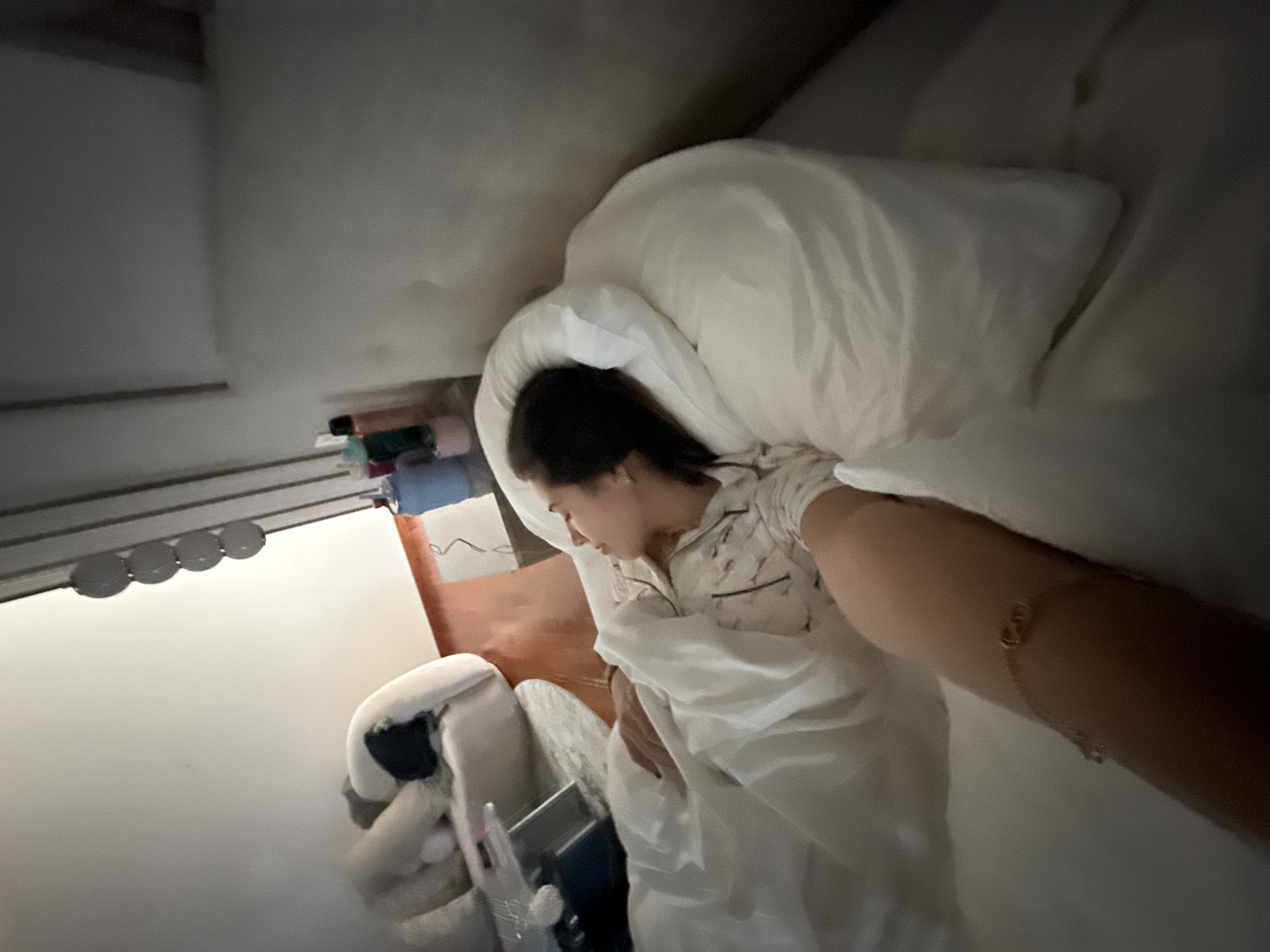 The image, captured from a high angle and rotated counter-clockwise, depicts a young female resting in a bright bedroom. She is lying on her side in a bed with white pillows and a white duvet or sheet, wearing light-colored pajamas with a subtle pattern. Her eyes are closed, indicating sleep or deep rest. Her right arm is extended towards the camera, adorned with a thin gold bracelet that features small charms, possibly "CD."
To the left of the bed, a white surface serves as a nightstand or shelf, holding several bottles or containers of various colors (blue, pink, brown). Below this, a white chair or stool has a dark object, possibly a bag or headphones, placed upon it, alongside a mobile phone on a stand. A horizontal strip of four circular light fixtures is mounted on the light-colored wall above the shelf. A glimpse of an orange-brown element, likely furniture, is visible further in the background.
The lighting in the room is soft and ambient, contributing to a calm and serene atmosphere. No specific time of day is clearly indicated, but it appears to be a peaceful moment. The provided context indicates the location as San Bernardino Tlaxcalancingo, Mexico, though no direct visual cues within the image confirm this. No legible text is visible.