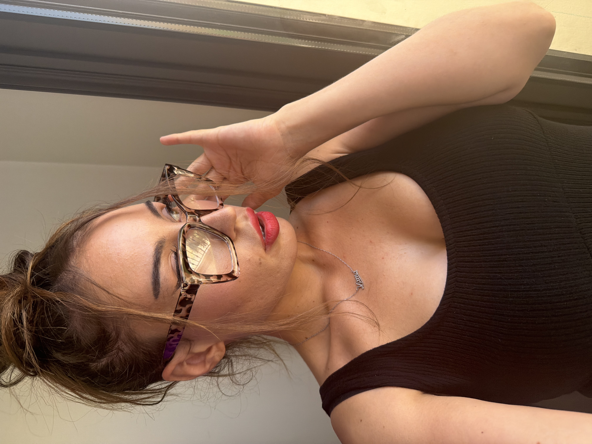 The image shows a medium close-up shot of a woman with brown hair pulled up in a messy bun. She is wearing large, leopard-print glasses and red lipstick. Her head is turned to the side, and she is looking out a window. Her right hand is raised to adjust her glasses. She is wearing a dark, ribbed tank top and a silver necklace that spells out "Mama." The background appears to be an interior setting near a window with natural light streaming in. The location of Tersefanou, Cyprus is not discernible from the image.