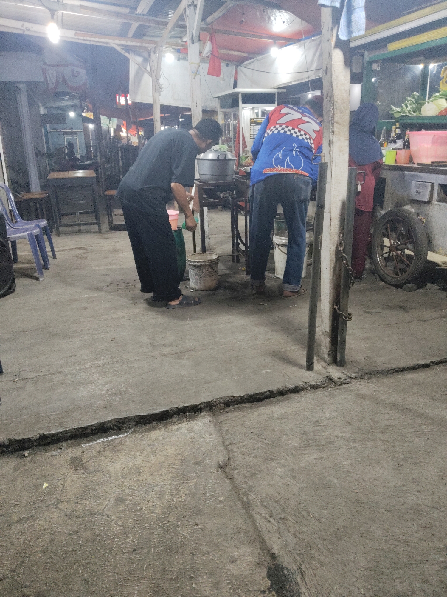 The image depicts an outdoor street food stall or small restaurant at night in Subang, Indonesia. The ground is concrete and shows signs of wear and tear, with a noticeable crack running across the foreground. In the background, there is a covered area with overhead lighting, likely part of the stall's structure.
Three individuals are visible. The person on the left, wearing a dark t-shirt and black pants, is bending over a large pot or cauldron, possibly preparing food. They are wearing dark sandals. To their right, a second person in a blue jacket with a checkered pattern and jeans is standing, facing away from the camera. Next to this person, on the far right, a third individual wearing a dark blue hijab and a maroon garment is also standing, facing away from the camera.
Various objects are present, including buckets, what appears to be a food cart or preparation counter, and a bicycle wheel mounted on its side in the far right background. The overall atmosphere is that of a busy, functional food stall operating in the evening.
