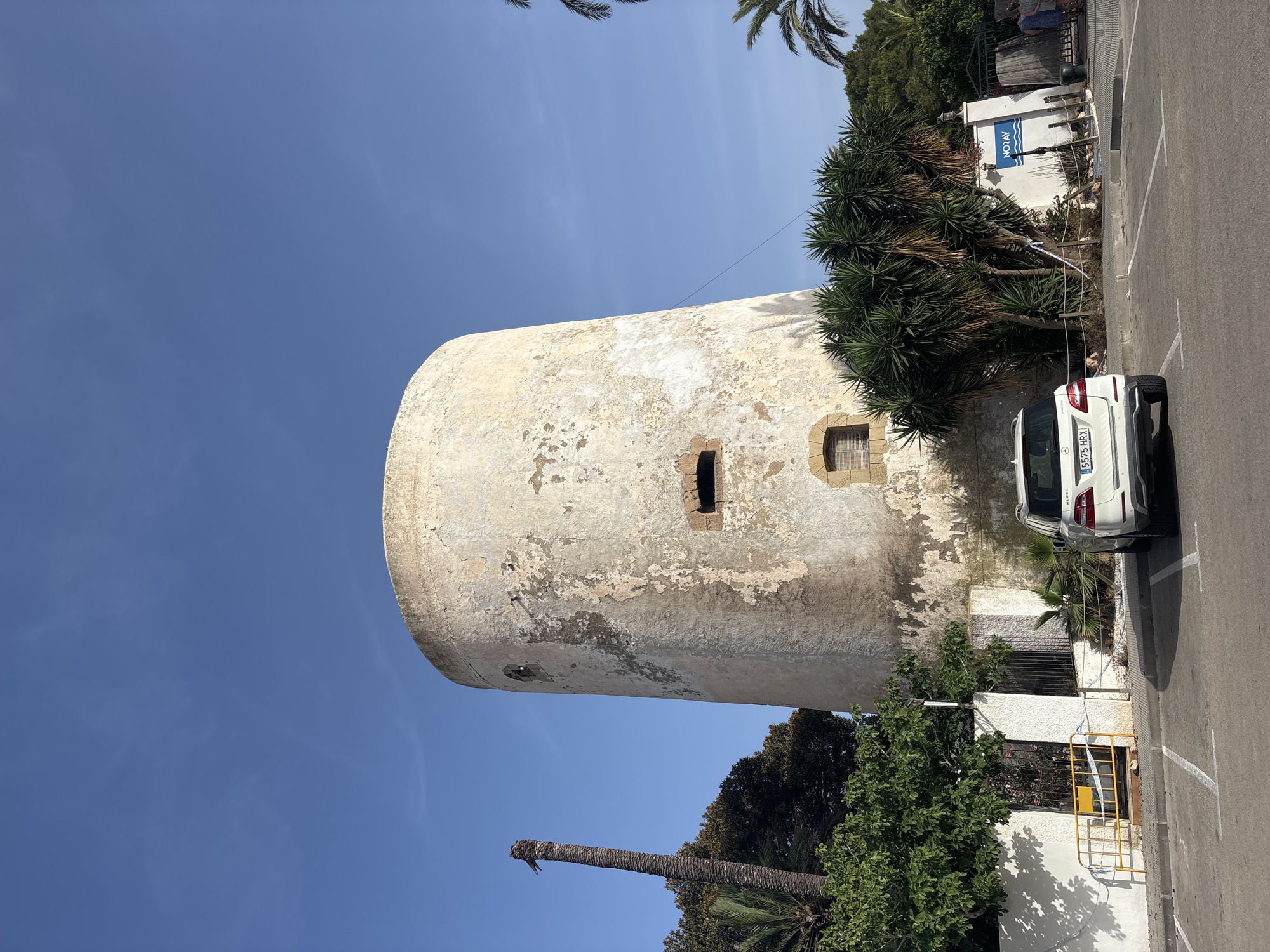 The image depicts a circular, weathered tower in Urbanización Cabo Roig, Spain. The tower is made of light-colored stone with visible patches of peeling plaster, revealing darker sections underneath. It has several small, dark window openings and a larger archway above them. A date stone is visible near the bottom.
Surrounding the tower, there is lush vegetation, including palm trees, yucca plants, and other green foliage. A white fence with yellow caution tape is present in front of the tower, with a yellow metal barrier visible behind it.
In the foreground, a white Mercedes-Benz SUV is parked in a paved lot marked with white parking lines. The car's license plate reads "5575 HRX". To the right of the tower, a white sign with blue lettering that says "NORAY" is mounted on a wall. A metal gate is visible behind the sign, with two women standing near it.
The sky is a clear, bright blue. To the left of the tower, a tall, dead palm tree stands with a bird perched on its top. The scene appears to be illuminated by natural daylight, casting shadows from the vegetation onto the ground and the tower.