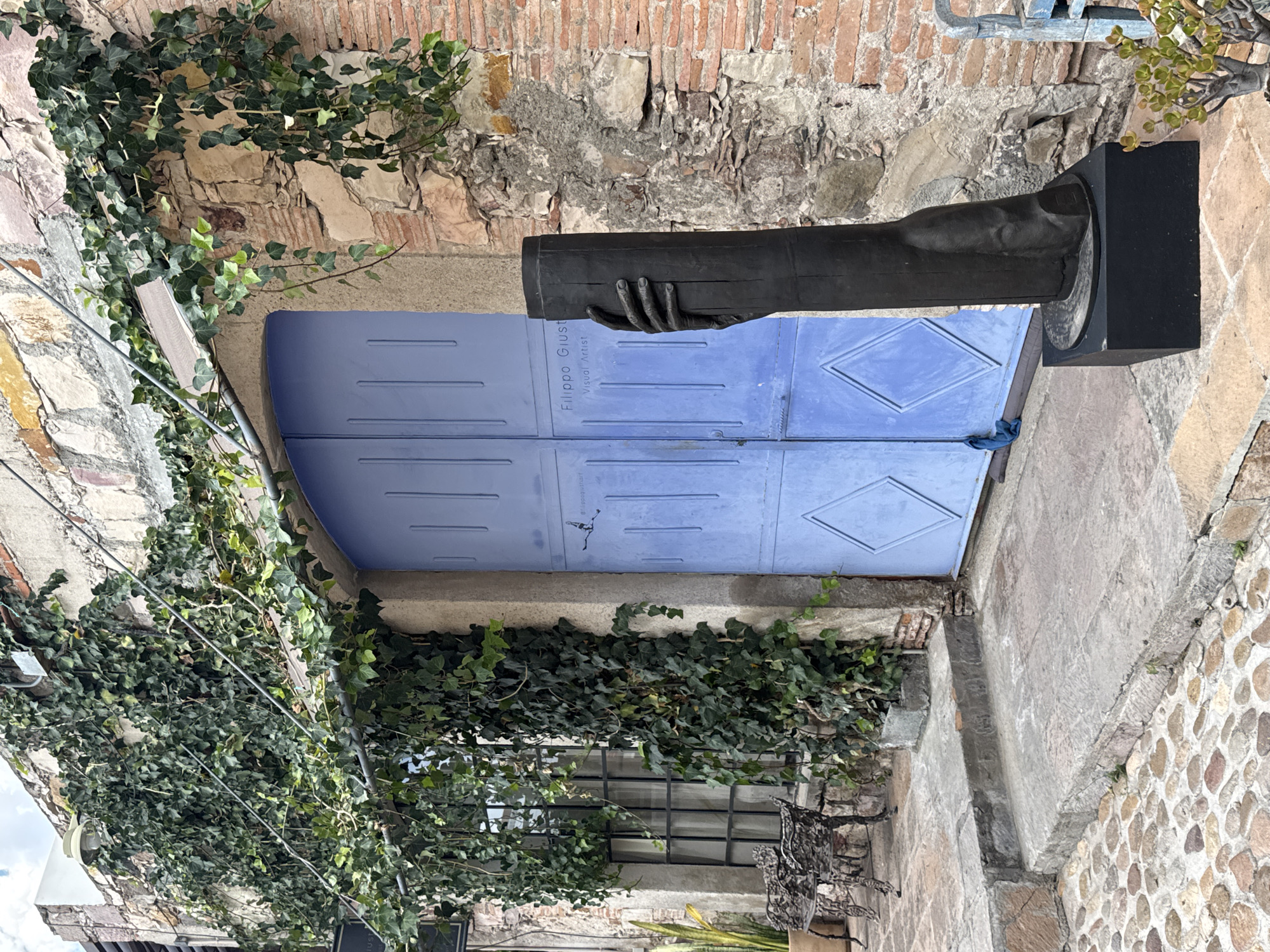 The image shows a building facade in San Miguel de Allende, Mexico. A blue arched double door is visible, with the inscription "Filippo Giusti, Visual Artist" on the door. A dark metal sculpture of a hand holding a cylinder is mounted on a black pedestal beside the door. The building exterior is constructed of stone and brick, with ivy growing on the walls and around a barred window below the door. The image appears to have been rotated 90 degrees.